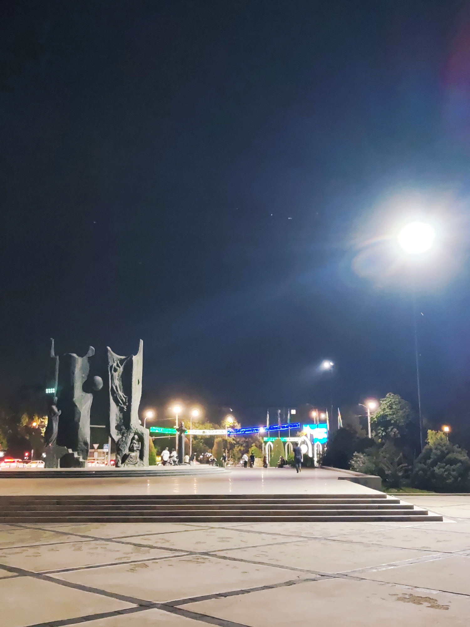 The image captures a nighttime view of the Amir Timur Square in Tashkent, Uzbekistan. In the foreground, a wide, paved plaza with a grid pattern is visible, leading up to a wide set of concrete stairs. Behind the stairs, a large, monumental sculpture dominates the scene. The sculpture appears to be made of stone or concrete and features abstract, human-like figures. One section of the sculpture shows a figure carved into its surface, possibly a relief.
Further back, beyond the sculpture, is a street with traffic lights and passing cars, indicating a city environment. Several lampposts illuminate the area, with a particularly bright streetlamp in the upper right corner causing lens flare. In the middle ground, there are several people walking and standing near the sculpture and the street. Some are silhouetted against the lights, while others are more visible. Trees line the background, and flags are visible on poles, suggesting a public square or park. The sky is dark, with a few faint stars visible, and the overall atmosphere is one of a well-lit urban public space at night.