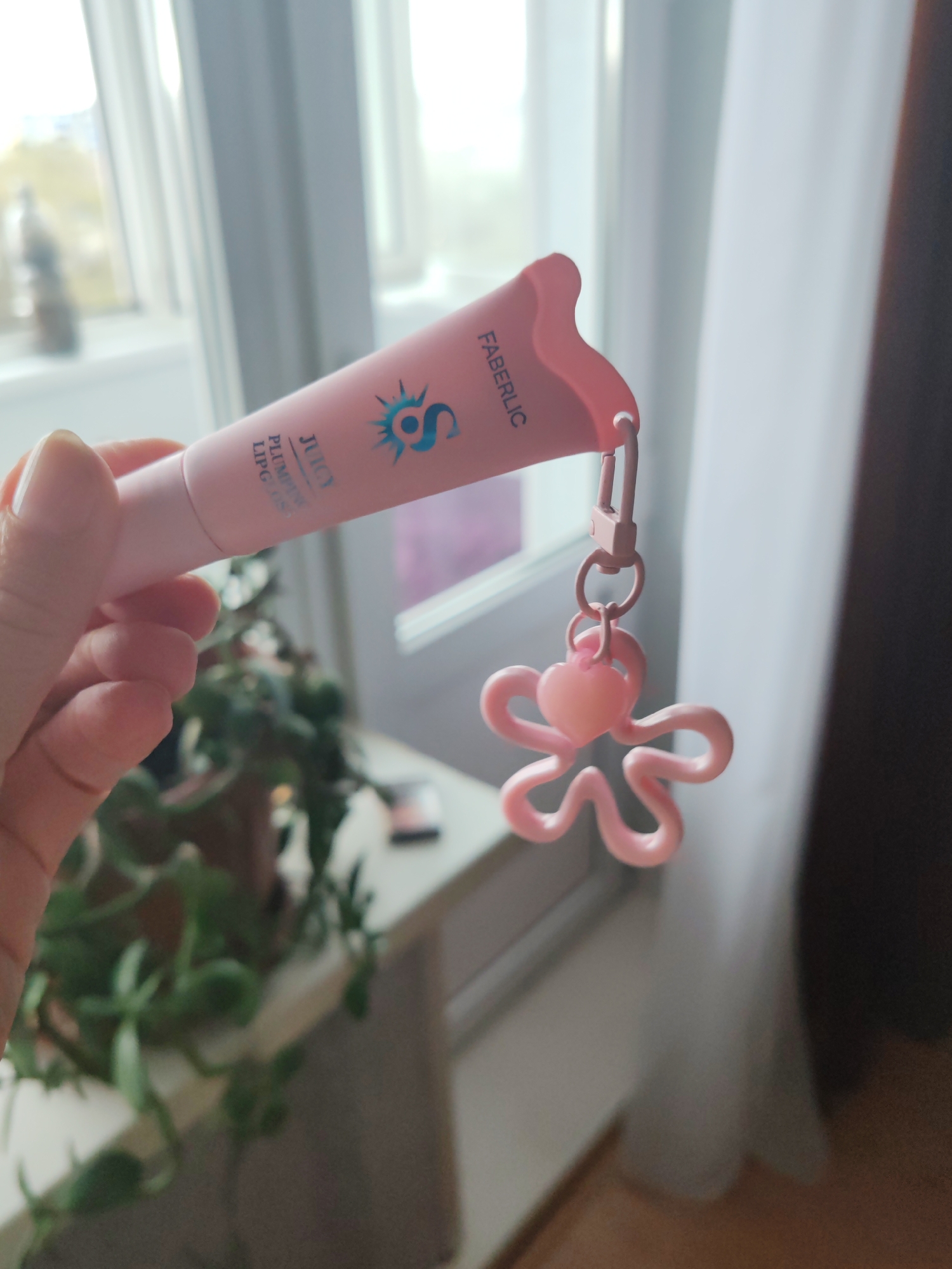 A hand holds a pink tube of Faberlic Juicy Plumping Lip Gloss with a pink flower charm attached. The background shows a window, a plant on a table, and part of a curtain. The location cannot be confirmed as Petropavlovsk-Kamchatsky, Russia without further visual or contextual information.