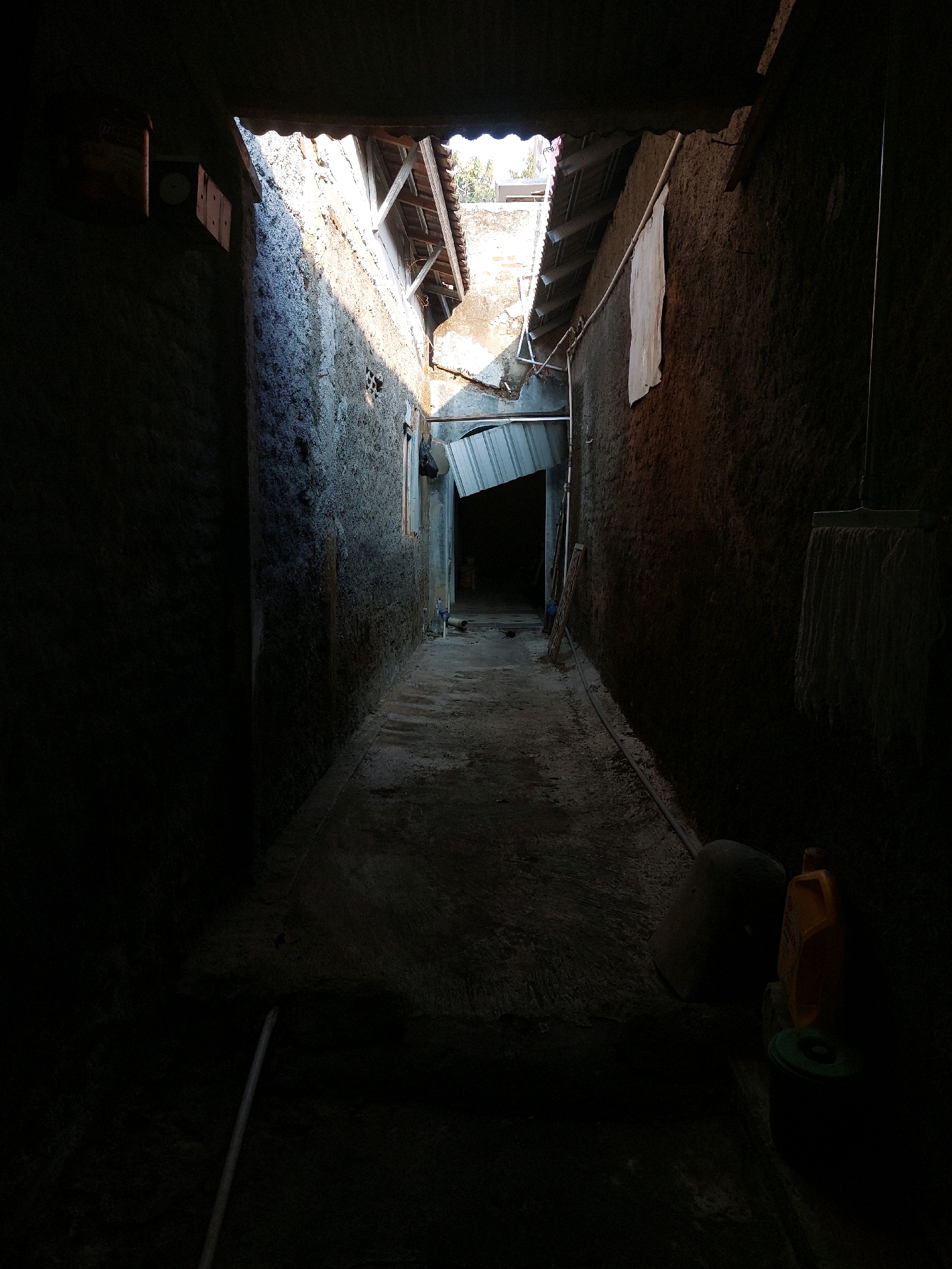The image depicts a narrow, dimly lit alleyway between two rough, concrete walls. The alley is covered by a dilapidated roof structure made of corrugated metal and wooden beams, with sunlight filtering through gaps and openings. In the foreground, a light-colored hose lies coiled on the ground. To the right, near the wall, a yellow plastic container, a large dark object, and a green plastic lid are visible. Further down the alley, a mop hangs from the wall. The ground is uneven and appears to be made of concrete or packed earth. The overall impression is of a neglected and somewhat dilapidated urban environment, likely a residential area in Selorejo, Indonesia, given the architectural style and the presence of everyday items.