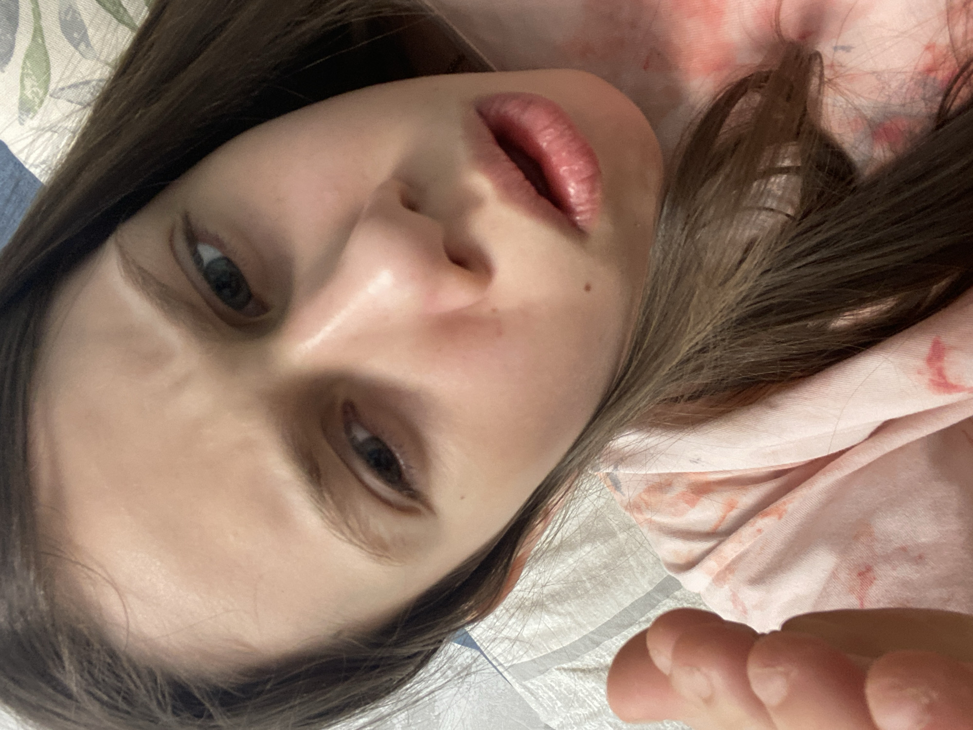 A close-up, angled selfie features a young person with fair skin, brown hair, and visible lips. The person is lying down on a white, textured surface, possibly bedding. The top part of the person is covered in a pink shirt. Parts of their fingers can be seen at the bottom of the image. The location in Izhevsk, Russia, is unknown and cannot be determined from the image.