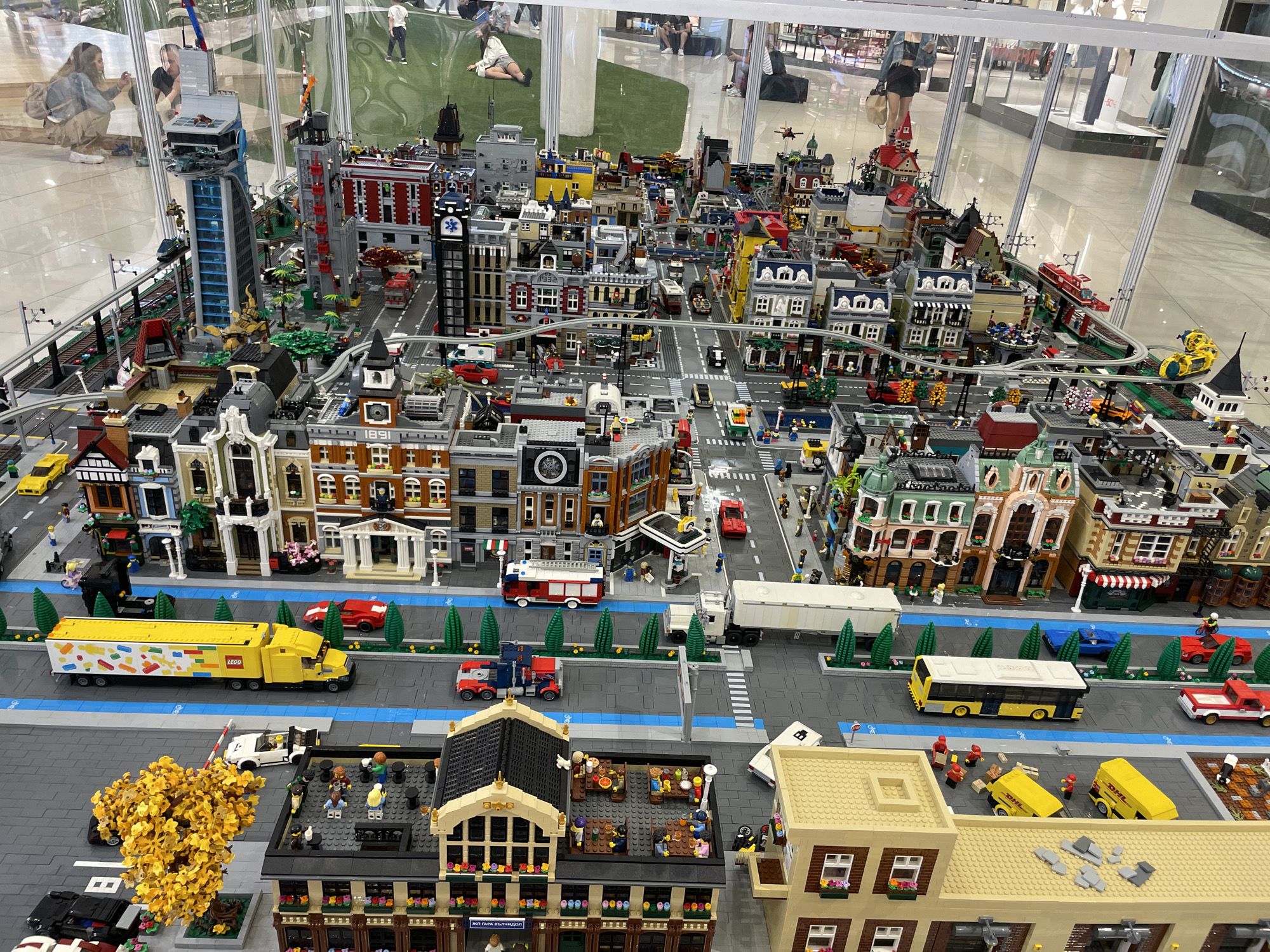 This is a detailed Lego model of a city, possibly representing a district in Sofia, Bulgaria, given the presence of what appears to be Cyrillic text on a building facade that reads "ЖИЛ. ГАРА ВЪЗРАЖДЕНЕ" which translates to "Residential Garage Vazrazhdane." The model features a complex urban landscape with numerous buildings of varying architectural styles, including residential blocks, commercial establishments, a fire station, and a tall office building with a distinctive blue and silver design.
Multiple Lego vehicles are integrated into the scene, such as a yellow Lego truck with colorful decals, a yellow bus, a red fire truck, red and blue Lego trucks, and various cars. Roads are laid out with white and blue lane markings, and streetlights are visible. Some buildings have Lego figures of people interacting, for example, gathered at outdoor seating areas with tables and chairs.
The model is situated within a large, transparent enclosure, suggesting it is an exhibition or display. In the background, through the enclosure, can be seen people observing the Lego city. The floor within the enclosure is tiled, and outside, there are elements that resemble a shopping mall environment with polished floors and reflective surfaces. In the background, there's a section of artificial green turf with some Lego figures in casual poses, further indicating a public exhibition space.