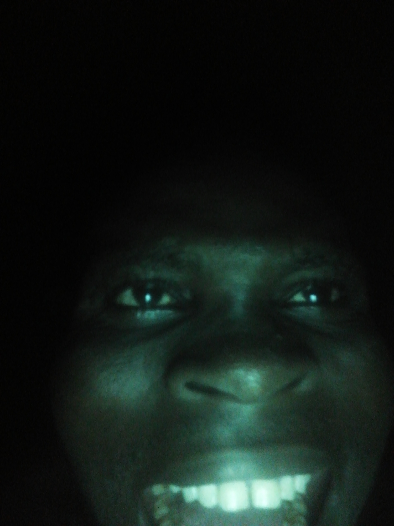 A close-up photograph shows a person's face in very low light conditions, with a noticeable greenish tint. The focus is primarily on the eyes, nose, and mouth, with the teeth visible in an open-mouthed expression. The surrounding environment is obscured by darkness. The location, Ikorodu, Nigeria, cannot be determined from this image.