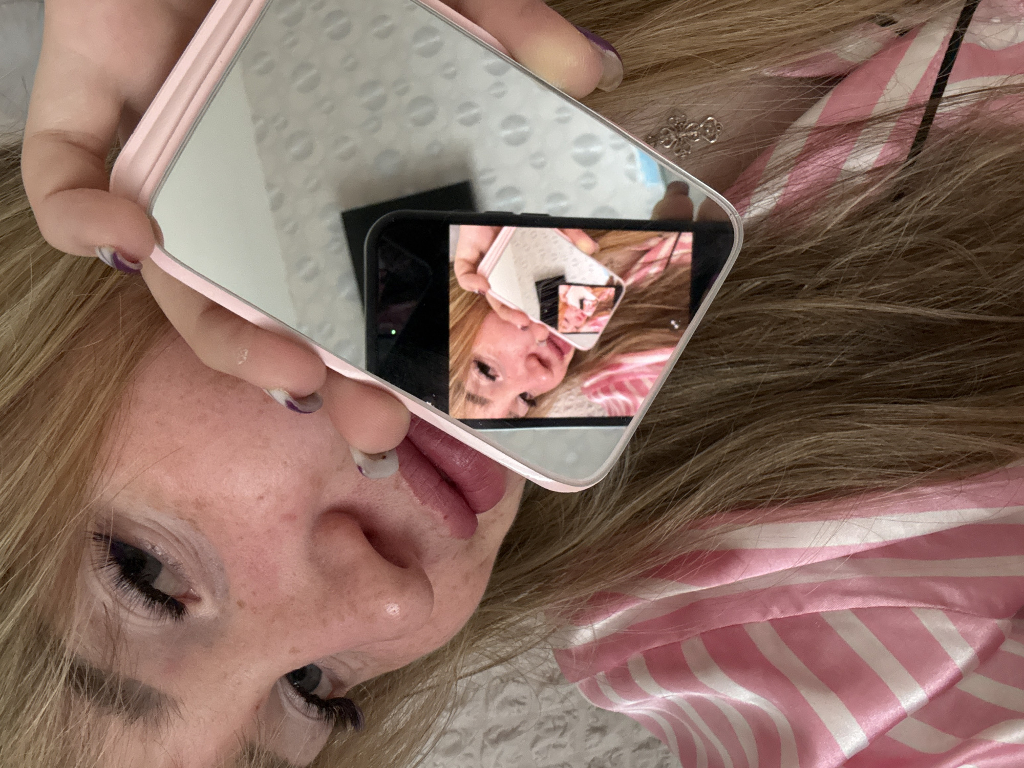 A close-up, high-angle shot captures a person, likely female, lying down with long blonde hair spread around her head. She has visible eye makeup, including long dark eyelashes, and her lips are painted with pink/mauve lipstick. One of her fingernails, visible on her left hand holding a phone, is painted with purple and white swirls, while another visible nail is white.
The central activity is the person taking a selfie using a smartphone encased in a pink, fully mirrored case. The reflection on this mirrored case prominently displays a black smartphone pointed towards the person, actively capturing the image. This creates a recursive "infinity mirror" effect, where the reflection shows the phone taking a picture of the person, and within that reflection, another image of the phone is visible, and so forth.
A silver-colored piece of jewelry, possibly a cross or an ornate design, is visible near the person's hair on the right side of the frame. The person is wearing a pink and white striped garment, which appears to be made of satin, possibly pajamas or a robe. The background visible in the mirrored reflection is a light-colored surface with a subtle, circular textured pattern. The person's expression is neutral as she looks towards the reflected camera. There is no visible text in the image. The scene is indoors, but specific time of day, weather, or visual cues confirming the given location of Марковский, Russia, are not present.