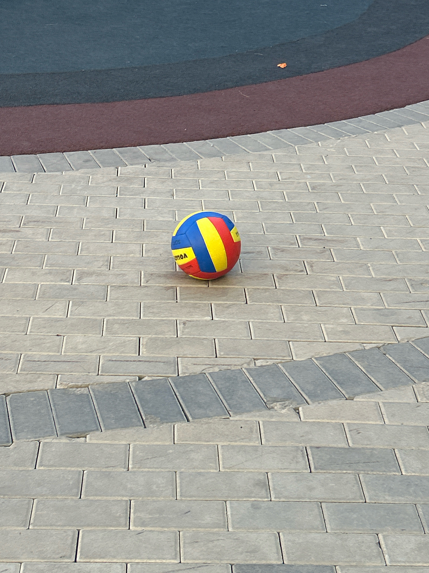 A vibrant volleyball, predominantly yellow with blue and red panels, rests on a paved walkway. The walkway is constructed from rectangular, light gray bricks laid in a running bond pattern, with a contrasting band of darker gray bricks forming a curved border. In the background, a section of a sports court is visible, featuring a dark blue surface, a thin maroon stripe, and a subtle transition to a black top layer. The scene appears to be outdoors on a bright, possibly sunny day, given the lighting and shadows. The image is likely captured in Astana, Kazakhstan, given the context provided, potentially at a public park or recreational area.