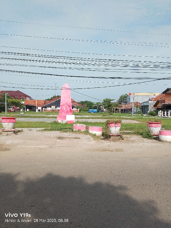 The image shows a pink and white obelisk monument in a small, open-air public square in Jatibarang, Indonesia. The monument is located at the center of the square, with several flower pots situated around it. The square is surrounded by buildings and trees. There are many electrical wires running across the sky. The ground is paved with asphalt. There are no people present in the image. The sky is cloudy, and the sun is not visible. The ground is covered in grass. A few trees and bushes are also visible. The image was taken on March 28, 2023 at 08:38 AM. The image was taken by Akhtar & Arvan. The phone model used to take the image is vivo Y19s.
