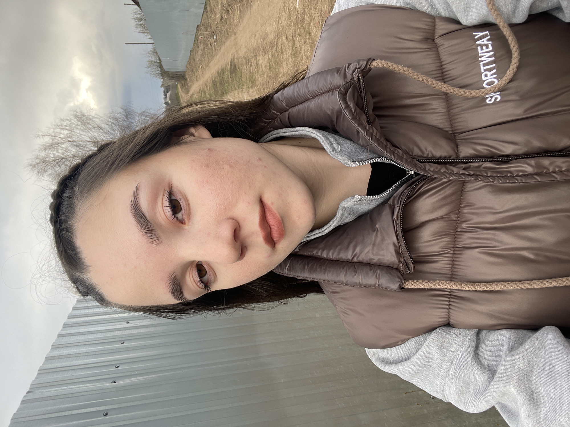 A close-up portrait features a young individual, likely female, posing outdoors. She has dark hair, partially styled with a braid along her hairline, and warm brown eyes with defined eyebrows. Her lips are naturally colored or lightly made up, and her skin shows some minor blemishes. She wears a brown or muted bronze puffer vest over a light grey hooded sweatshirt, with a black garment visible underneath. The vest has a braided drawstring around the collar.
The word "SPORTWEAY" (possibly a stylized "SPORTWEAR") is embroidered in white on the right chest area of her vest.
The person is looking towards the viewer with a neutral or slightly contemplative expression, suggesting a selfie or a posed photograph.
The background indicates a rural or semi-rural outdoor setting, possibly in Ольховка, Russia. To the left, a corrugated metal fence runs horizontally. To the right and behind her, a dirt path or road recedes into the distance. Bare trees, indicative of late winter or early spring, stand against an overcast, cloudy sky. The lighting suggests it is daytime, and the attire implies cool weather.