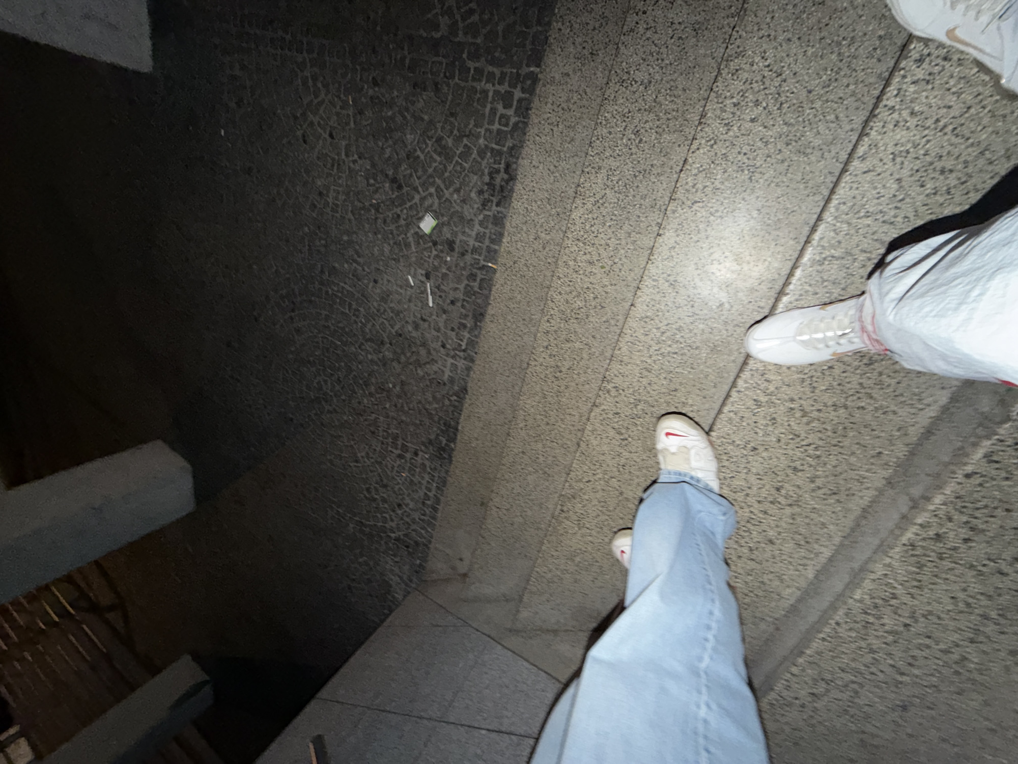 The image displays an outdoor, low-light urban scene in Vila Real, Portugal. The perspective is downward, capturing the lower bodies of two individuals and their immediate surroundings.
Subject A, in the immediate foreground, wears light blue denim jeans and white sneakers with prominent red accents (likely Nike swooshes). Their feet are positioned on a multi-tiered staircase constructed from light grey, aggregate concrete or stone. Specifically, the right foot is on a lower step, while the left foot is on a higher step.
Subject B, partially visible to the right of Subject A, wears light-colored pants and white sneakers with light brown or beige accents on the laces and sole. Subject B's visible right foot is positioned on the same horizontal plane as Subject A's lower foot, adjacent to the staircase.
Beyond the staircase, the ground transitions to dark, tessellated Portuguese pavement, characterized by intricate, interlocking patterns of small stones. Scattered on this pavement are several white cigarette butts and one small, light green rectangular object. In the background, two light-colored, rectangular architectural columns or pillars are visible, rising from the pavement. The overall environment is dimly lit.