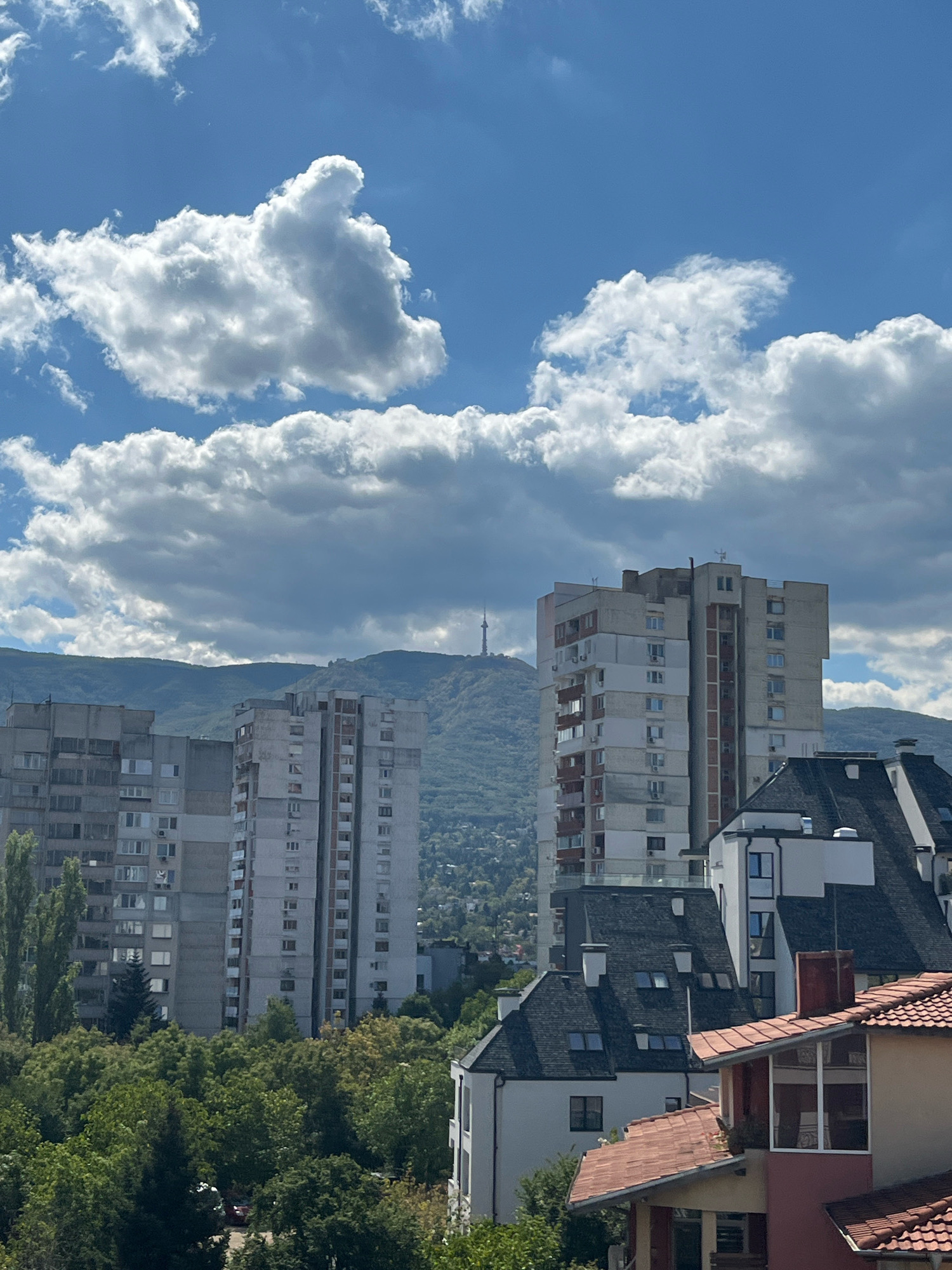 This is a vertical, outdoor, daytime photograph of the skyline of Sofia, Bulgaria, focusing on the residential areas of Mladost district and the Vitosha Mountain in the background.
In the foreground, several modern residential buildings with red-tiled roofs are visible. These structures are characterized by their multi-story design, varied window placements, and some architectural details like dormer windows.
Behind these, taller, older apartment blocks stand prominently. These buildings are constructed with concrete and feature numerous uniform windows arranged in vertical columns, typical of Soviet-era architecture. These are likely located in Mladost.
Further in the background, the lush green slopes of Vitosha Mountain rise, creating a natural backdrop to the urban landscape. Perched atop the mountain is a telecommunication tower, a recognizable landmark of Sofia. The sky above is a vibrant blue, dotted with fluffy white cumulus clouds, suggesting fair weather. The scene is devoid of any people or vehicles. The image captures a panoramic view of the city's transition from urban development to its natural mountainous surroundings.