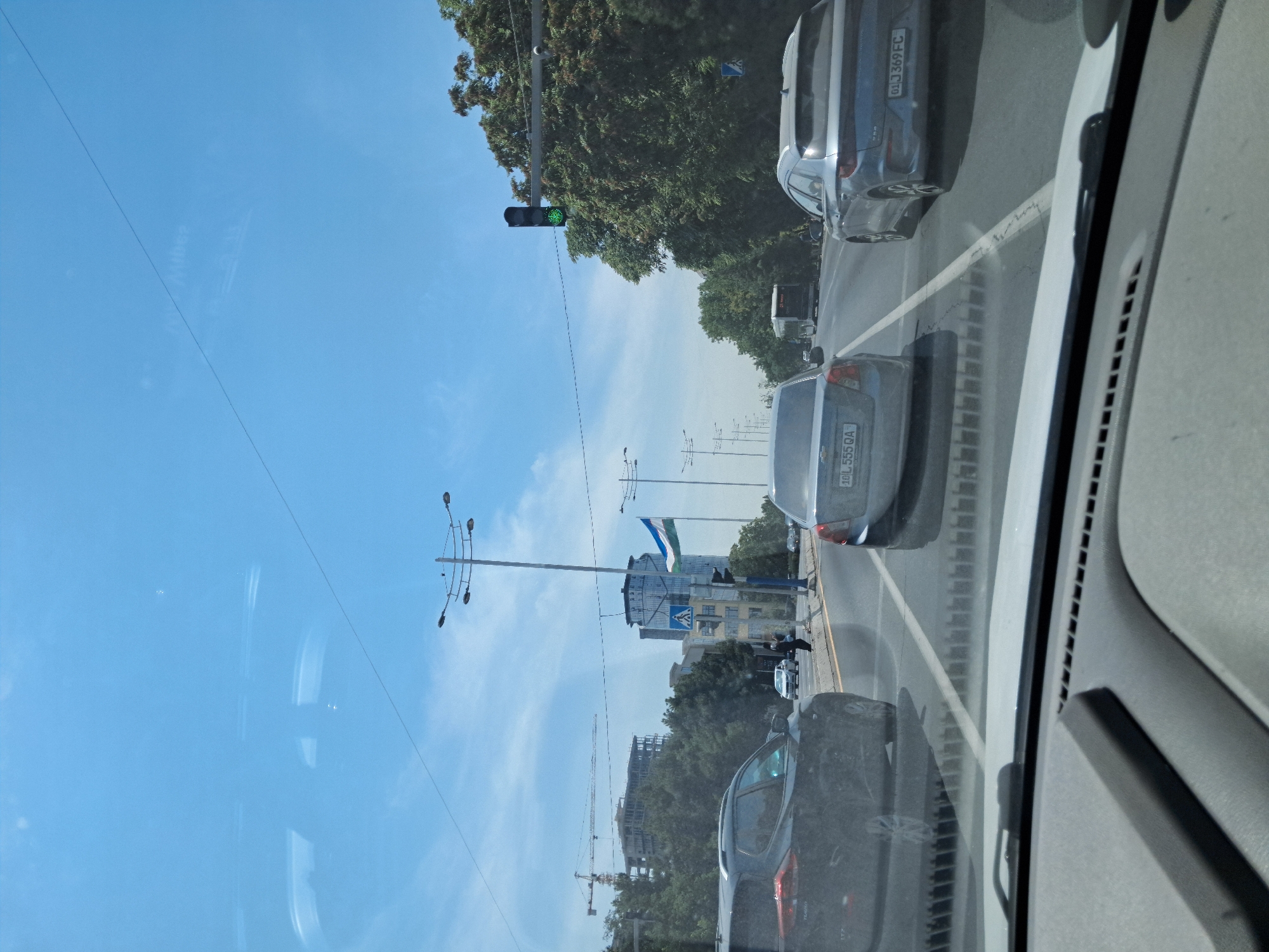 The image depicts a view from inside a vehicle traveling on a multi-lane road in Tashkent, Uzbekistan, on a clear, sunny day. The foreground shows the car's dashboard and windshield, with reflections visible on the glass. In the mid-ground, several cars are driving on the road, including a dark SUV on the left, a silver sedan in the center, and a light blue sedan on the right. The central sedan has a license plate that reads "10 L 555 QA," and the blue sedan has a plate reading "01 J 369 FC." Pedestrians are visible on a sidewalk to the right, near a traffic light which is currently green. Further back, several tall buildings and construction cranes indicate an urban environment. A large tree-lined area is visible on the right side of the road, and a flag of Uzbekistan is prominently displayed on a flagpole. The sky is a bright, cloudless blue, with faint reflections of the car's interior on the windshield. The scene appears to be a typical urban commute in Tashkent, possibly along a major thoroughfare like Amir Timur Street, given the presence of modern buildings and traffic infrastructure.