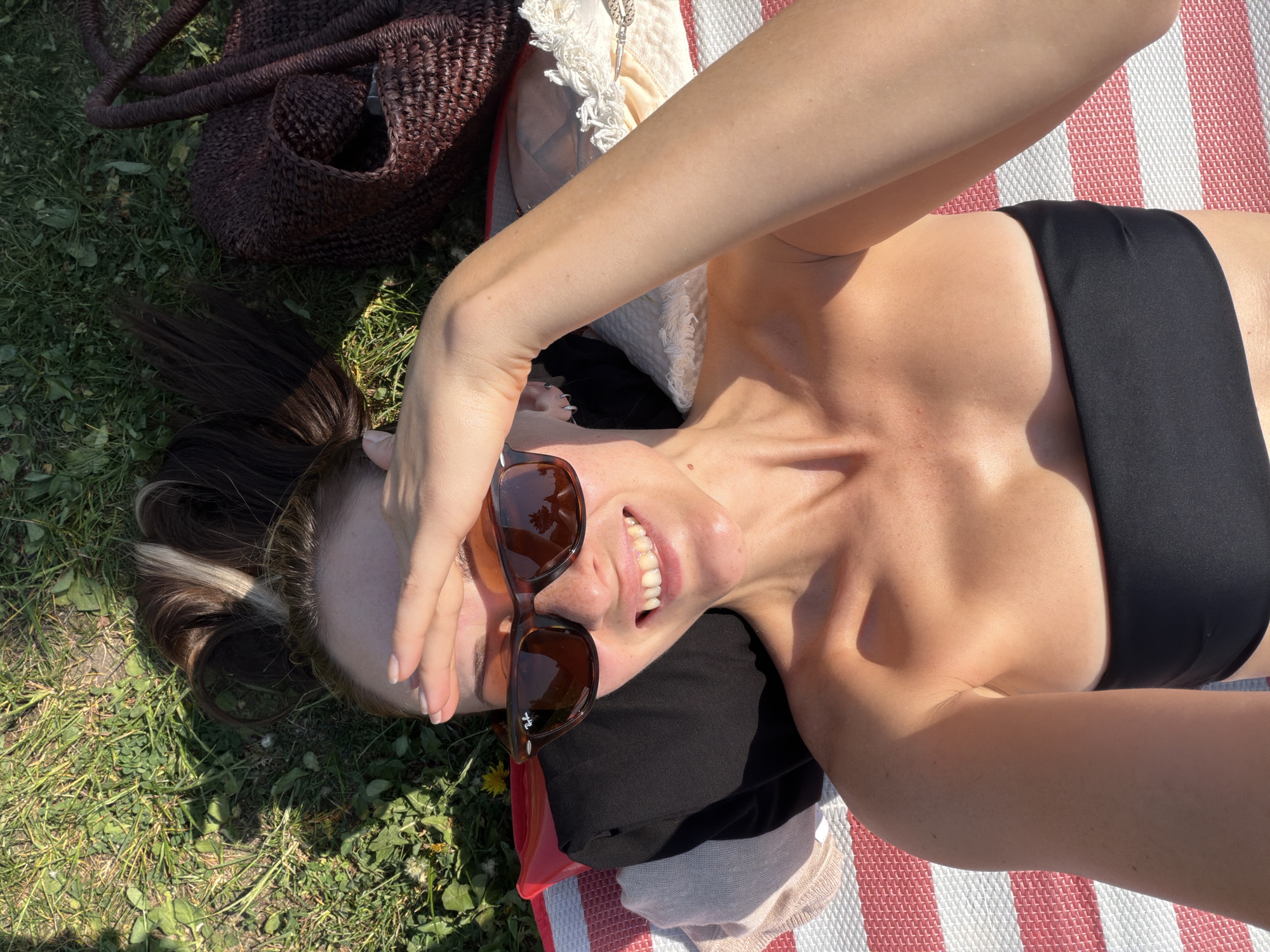 A woman is lying on her back on a striped blanket, which is partially on grass, possibly in a park. She is wearing a black swimsuit top and sunglasses, with her right arm raised, hand partially covering her face. A brown woven bag is visible near her head on the grass. Her hair is brown with a blonde streak and is tied back. There is a black garment under her head and a fringed cream colored cushion next to it. The location is not specified as Khimki, Russia.