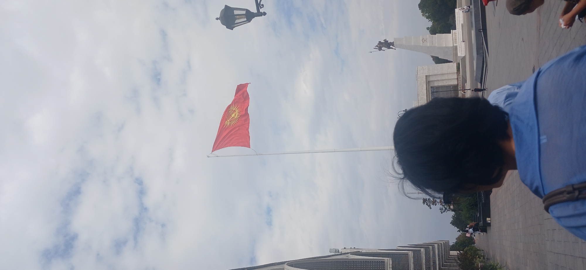 The image depicts a view of Ala-Too Square in Bishkek, Kyrgyzstan, on a cloudy day. In the foreground, the back of a person wearing a blue shirt is visible, facing away from the camera. Behind this person, a flag of Kyrgyzstan, red with a yellow sun in the center, is flying on a tall flagpole. Further in the background, a monument with a statue of a warrior on horseback is visible. The square also features a large, modern building with arched windows on the left side of the frame. The ground is paved with grey stone. The overall scene suggests a public gathering or a tourist location.