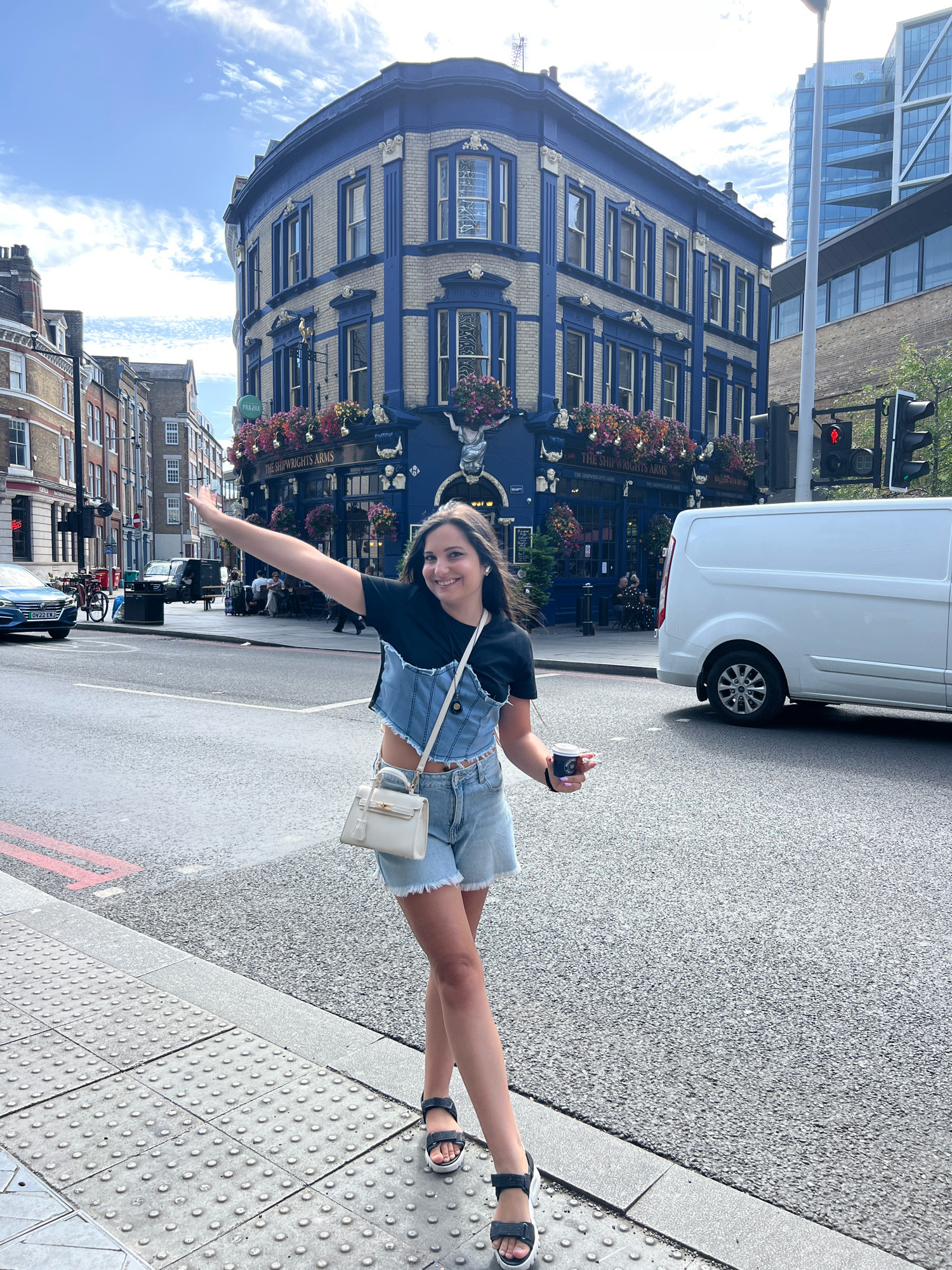 A young woman stands on a sidewalk in London, United Kingdom, with a building in the background. The woman is smiling and waving with her left arm outstretched. She is wearing a black t-shirt, a denim crop top, denim shorts, and black sandals. She is also carrying a small white handbag and a coffee cup. The building in the background is blue with white trim and has flowers on its balconies. The street is paved and there are traffic lights and a white van in the background.