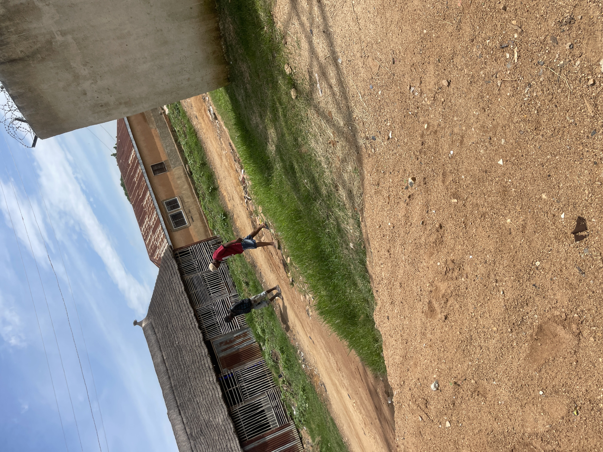 The image depicts an outdoor scene with two individuals walking on a dirt path. To the left, there is a building with a thatched roof and corrugated metal siding, which appears to be in a state of disrepair. Adjacent to it is a building with a rusty, corrugated metal roof. In the foreground, a sandy area is visible, with some dry grass and scattered debris. To the right, a portion of a concrete structure, possibly a wall, is present, topped with barbed wire. The sky is blue with scattered clouds. The environment suggests a semi-urban or rural setting, consistent with areas in Jos, Nigeria, characterized by traditional and modern architectural styles and unpaved pathways. The lighting indicates it is daytime, likely under a bright sun.