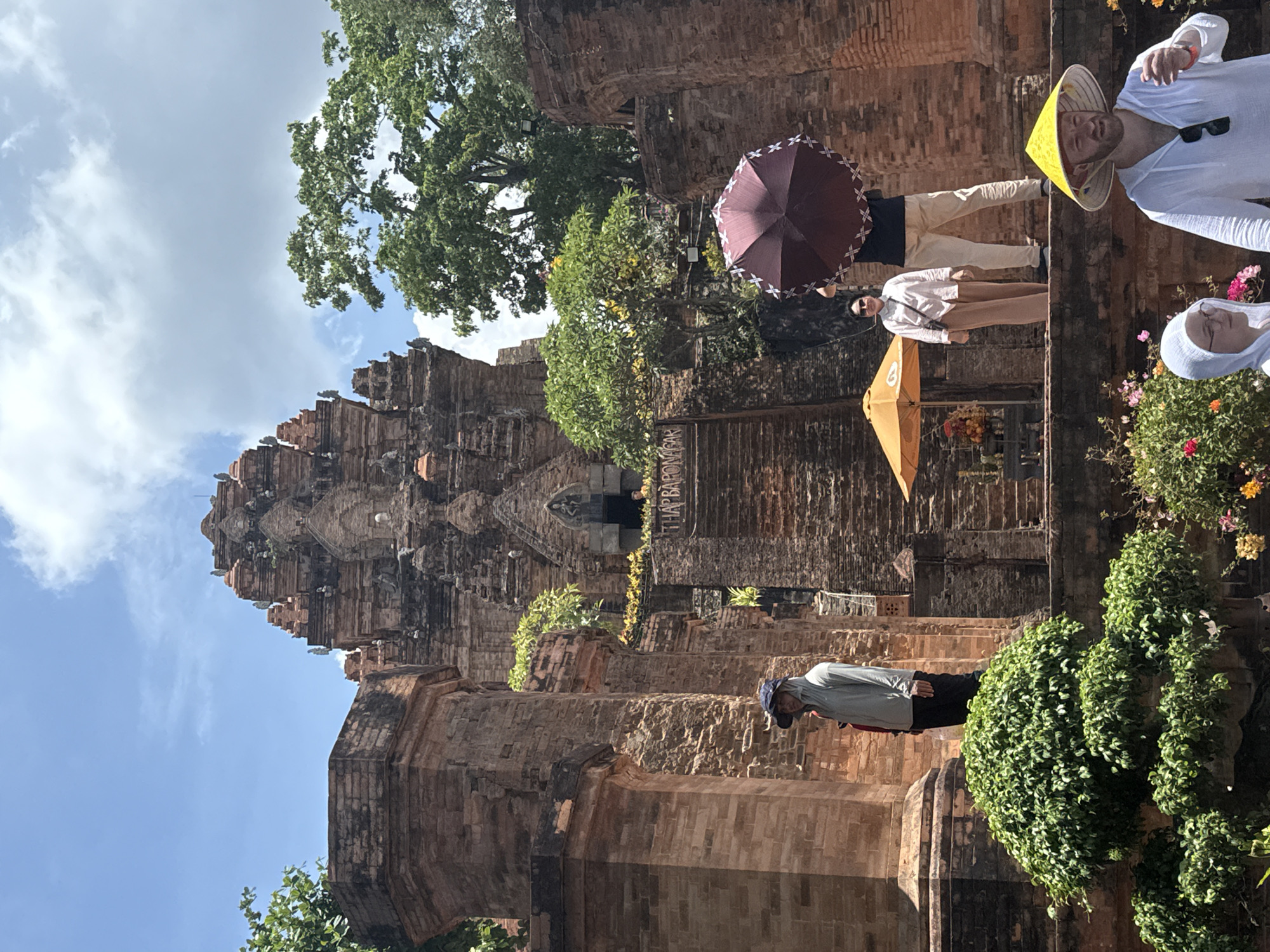 The image depicts a brick temple tower in the city of Bac Nha Trang Ward, Vietnam. Several people are present; one is wearing a yellow conical hat and a white shirt and sunglasses, while another is wearing a headscarf. The temple is adorned with greenery. There are multiple umbrellas.