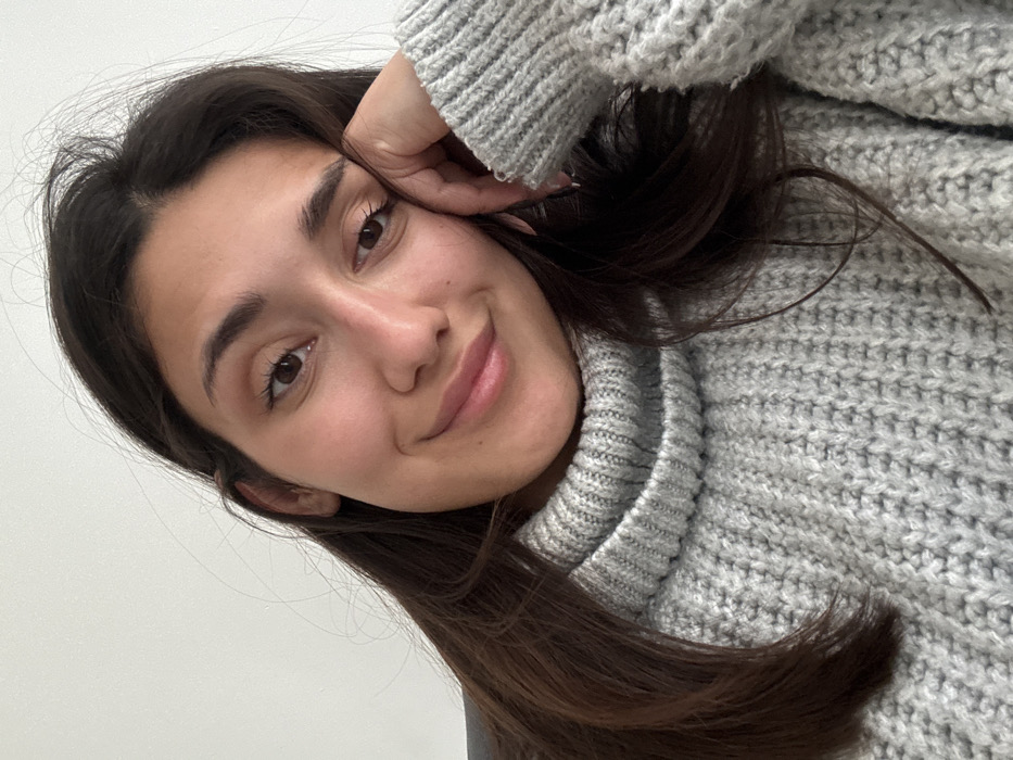 A young woman with dark hair and brown eyes gazes at the viewer with a gentle smile. She is wearing a chunky, light gray knit sweater with a turtleneck. Her head is tilted, resting lightly on her hand. The background is a plain white wall. She has smooth skin and her eyebrows are well-defined. The overall impression is one of casual comfort and warmth.