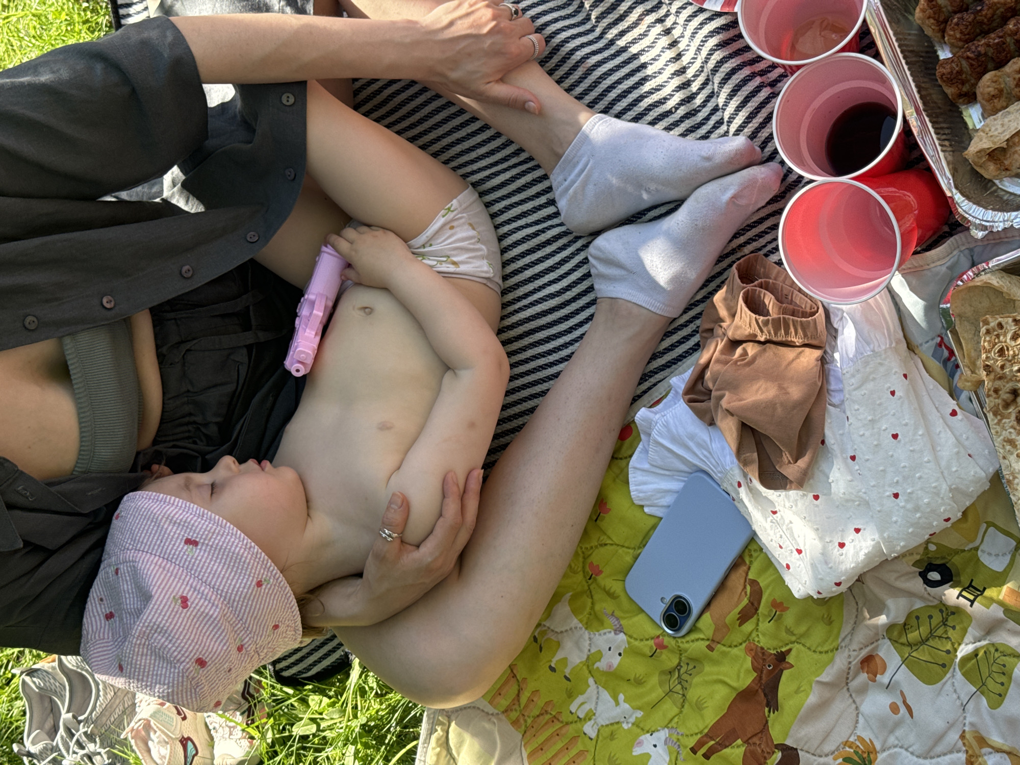 A photograph shows a woman and child reclining on a picnic blanket outdoors, likely on a sunny day. The child, wearing a cherry-print hat and light-colored diaper, is asleep in the woman's arms, holding a pink toy. The woman is wearing a button-down shirt and has rings on her fingers. The picnic blanket is striped in one section and patterned with green, yellow, and goat designs in another. Beside them, there are red plastic cups, a phone, some clothing, and food items in a foil container. The setting appears to be a park, with grass visible at the edges of the blanket. The location is not in Moscow, Russia.