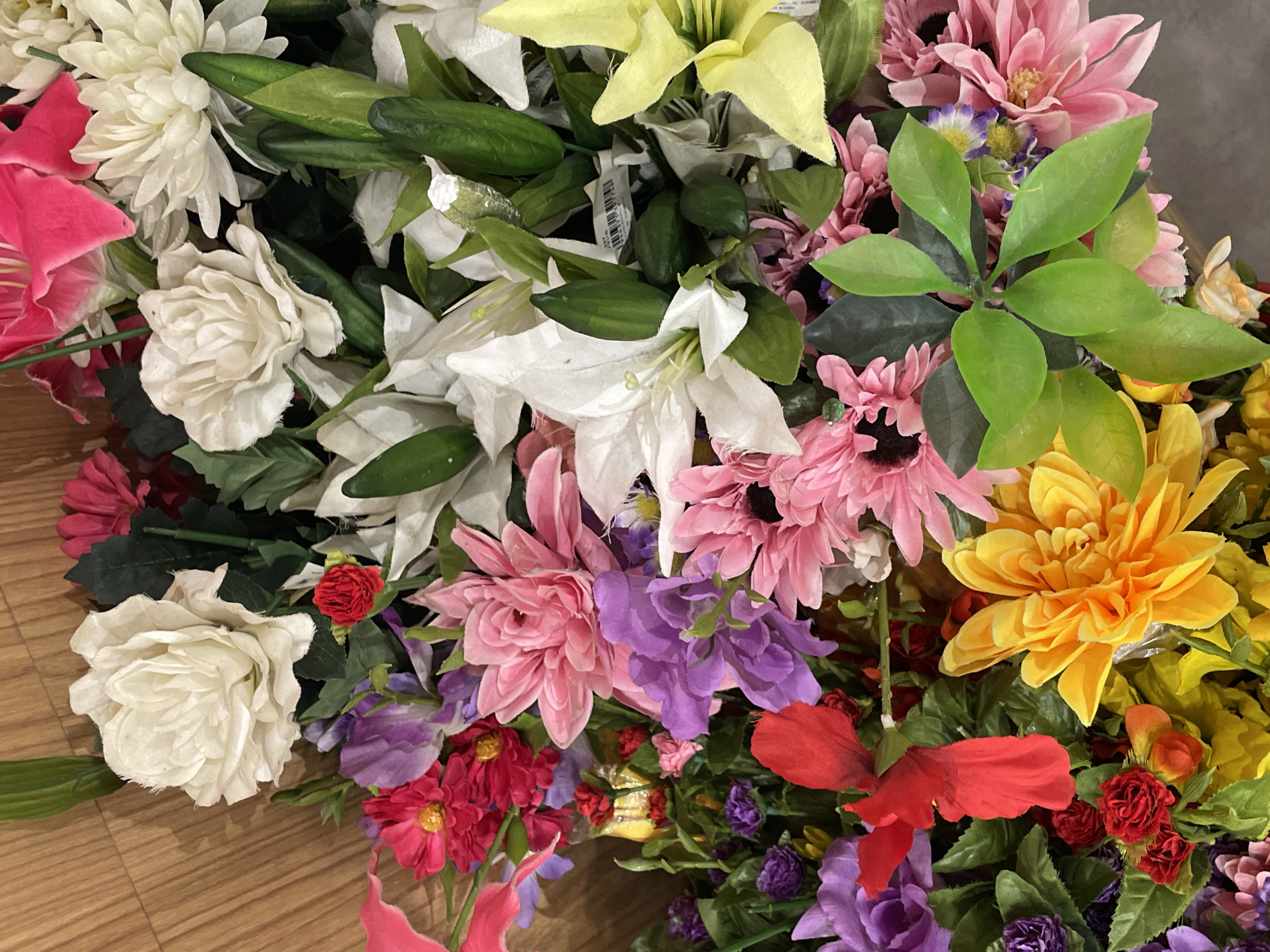 A close-up, overhead shot of a vibrant, artificial flower arrangement is presented. The bouquet features a diverse array of colorful flowers, including white roses, pink and yellow dahlias, purple irises, red hibiscus, and smaller red roses. Green foliage is interspersed throughout the arrangement. The flowers are densely packed together, creating a full and textured display. The background consists of a wooden surface, likely a table or shelf, and hints of a floral shop environment. The image does not depict any people or specific actions, and it is not possible to determine the location within Davao City, Philippines from the provided visual information.