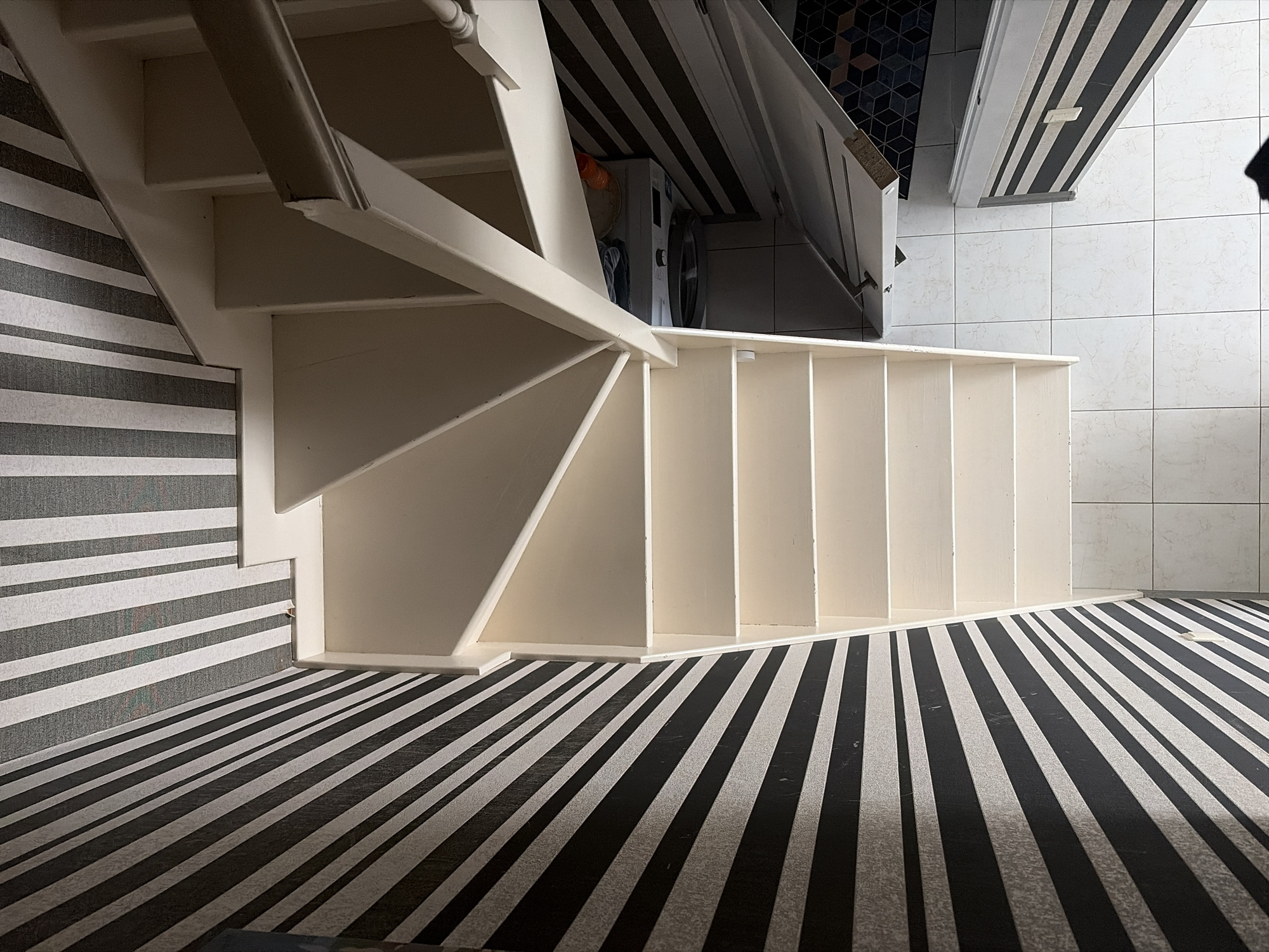 An interior residential space in Sochi, Russia, is depicted from an elevated, overhead perspective, looking down a winding staircase. The image is presented in a portrait orientation, rotated approximately 90 degrees clockwise.
The central feature is an off-white or cream-colored wooden staircase, comprising several flights and angled landings, indicating a U-shaped or L-shaped design. A white wooden handrail and balusters are visible along the open side of the staircase, particularly in the upper and middle sections.
The surrounding walls are covered in distinct black and white striped wallpaper. The wall to the left (bottom in a re-oriented view) displays diagonal stripes that appear to converge towards the lower part of the image. The wall on the upper right (top in a re-oriented view) features vertical black and white stripes.
Beneath the upper section of the staircase, a compact utility area is visible. It contains a white front-loading washing machine with laundry baskets or similar items stacked on its top. To the right of the washing machine, a white cabinet door, detailed with black and white horizontal stripes and equipped with two silver handles, is slightly ajar. Behind this door, a darker floor with a geometric, tessellated pattern is discernible.
The main floor area, situated at the base of the staircase, is covered with large, square white ceramic tiles that exhibit a subtle, light grey mottled pattern. The lighting appears even, suggesting ambient illumination without strong, directional light sources. No individuals or active interactions are present in the scene.