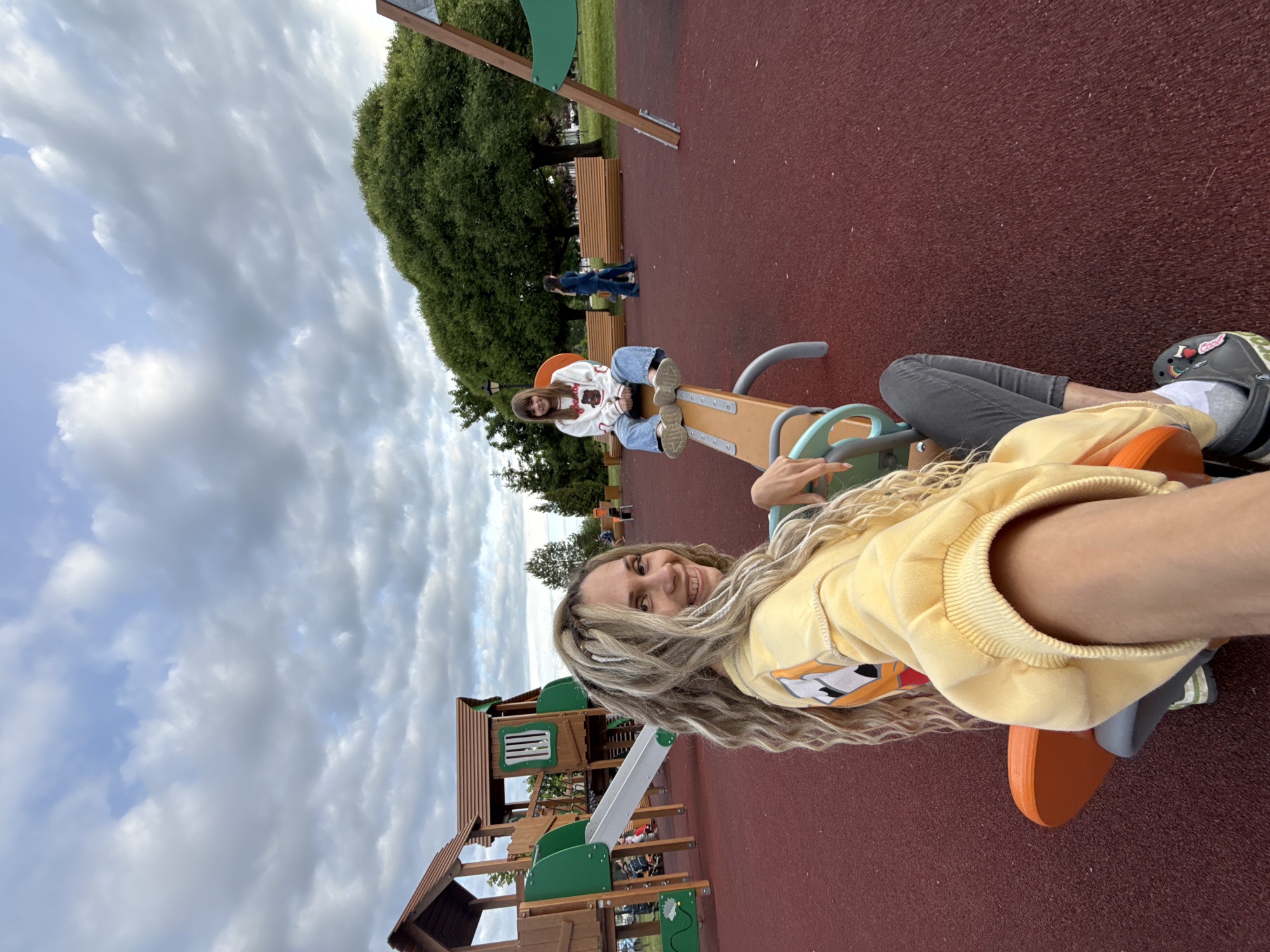 The image captures two women enjoying a playground on an overcast day in Заволжье, Russia. In the foreground, a woman with long, light blonde, curly hair (possibly dreadlocks or braids) is taking a selfie, smiling broadly at the camera. She is wearing a yellow hooded sweatshirt with an orange and white graphic, and dark grey pants. Her grey Croc shoe, visible in the lower right, features a charm that reads "I ❤️ God".
Behind her, another woman is seated on the opposite side of a two-person seesaw, which is orange and light blue/green. She wears a white patterned long-sleeved top, blue jeans, and sneakers, looking casually towards the camera. Further in the background, a third person in dark clothing walks past wooden benches.
The setting is an outdoor playground with a distinct red, textured rubber safety surface. In the background, a large wooden playground structure with a slide and climbing frames is visible. Tall, lush green trees frame the play area. The sky above is cloudy, with large patches of grey clouds, suggesting an overcast daytime. The overall mood appears relaxed and joyful.