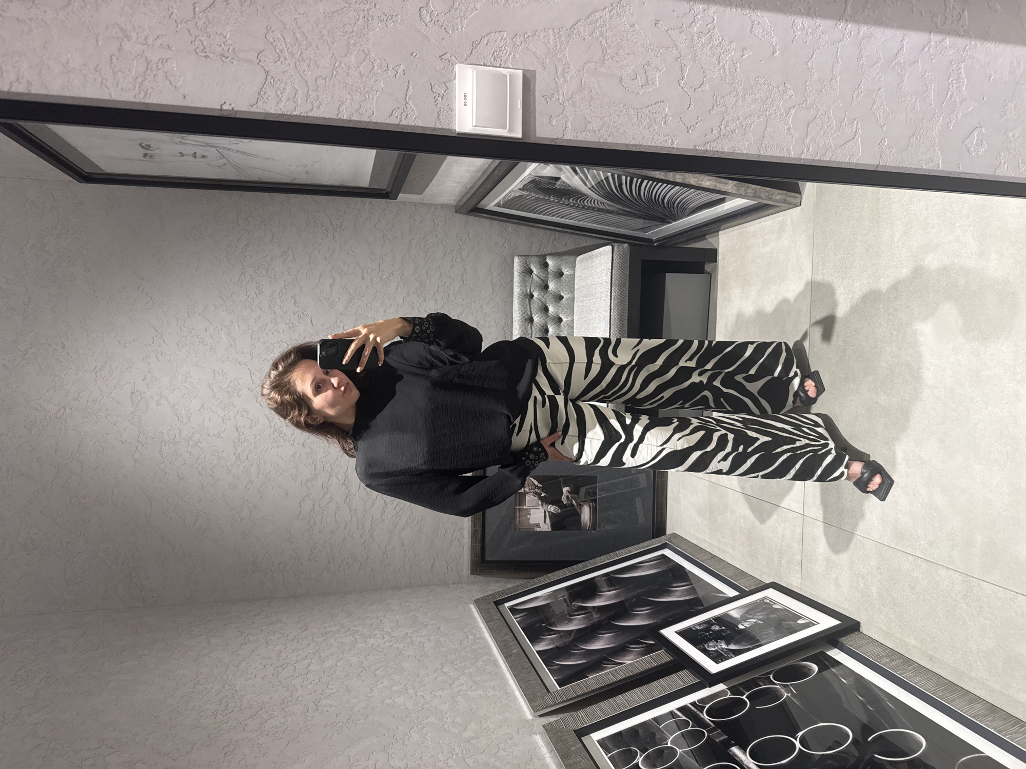 A woman takes a selfie in what appears to be a luxury interior. The woman is wearing a black top with details on the sleeves and zebra-striped pants with black heels. She is holding a smartphone. The environment includes decorative framed artwork on the wall, a padded chair and a light switch. The location is potentially in Dubai, United Arab Emirates.