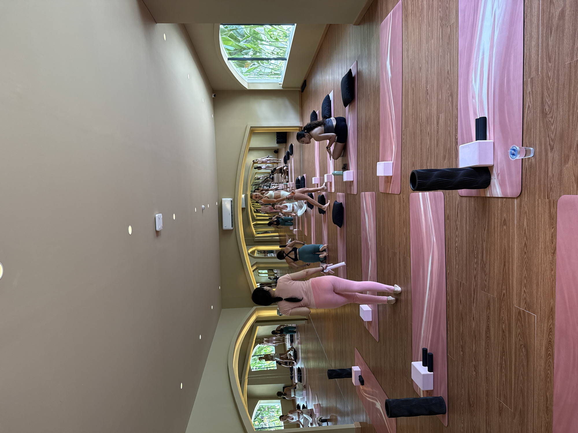 The image depicts an indoor yoga studio in Kuta, Indonesia, with wooden floors and large mirrored walls reflecting the scene. Several women are practicing yoga on pink yoga mats arranged in rows. A woman in pink leggings and a pink top, presumed to be an instructor, stands facing away from the camera, holding a white object. Other women are in various yoga poses, some seated, some standing, and some kneeling. Yoga blocks and foam rollers are visible on some of the mats. The room is well-lit, with recessed ceiling lights and natural light filtering in from arched windows on the right side of the image, which offer a view of lush greenery. The overall atmosphere suggests a professional and serene environment for physical activity.