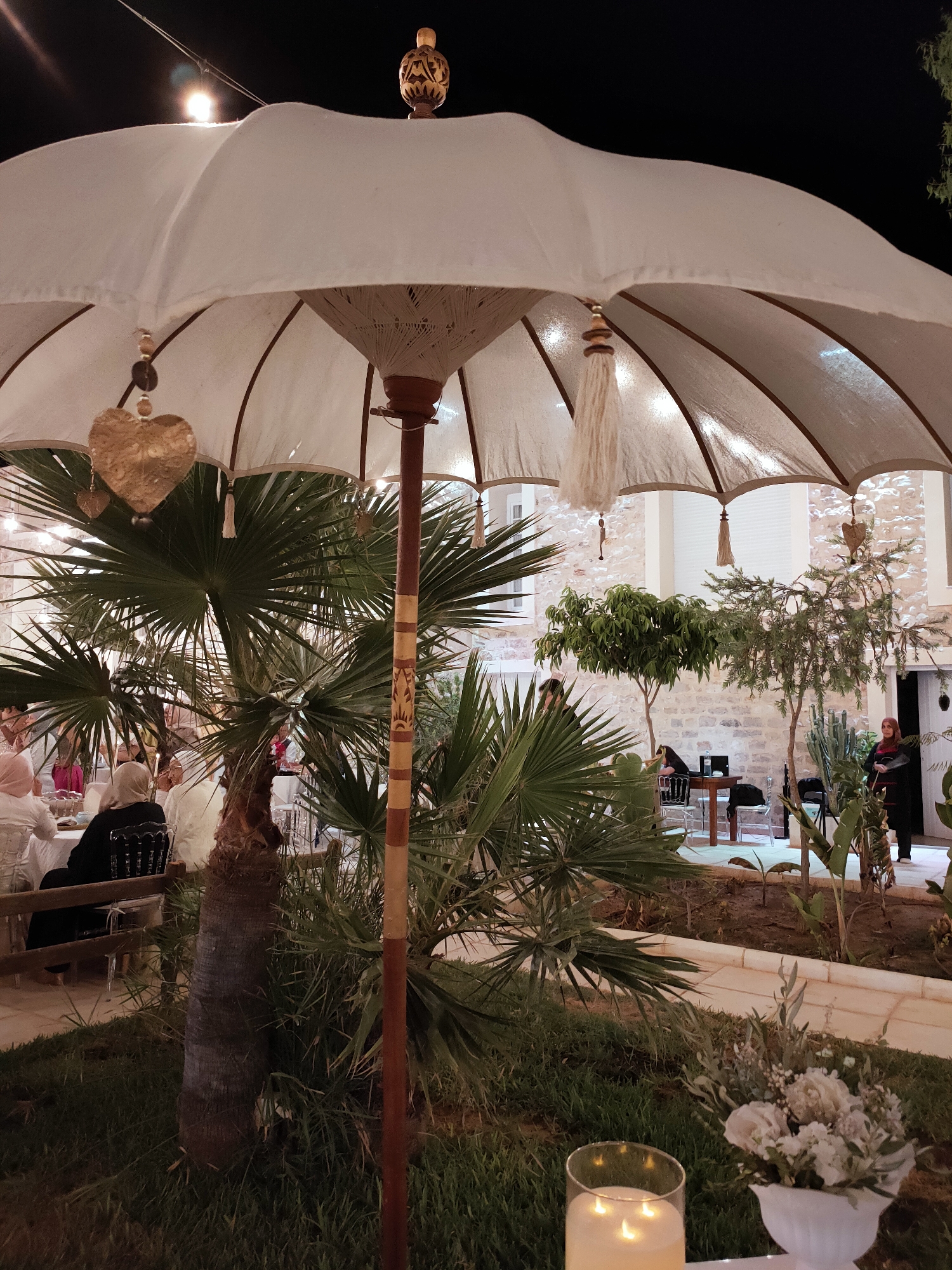 An outdoor evening scene in Cebalat, Tunisia, features a large, white umbrella with wooden accents, adorned with a gold heart charm and tassel. Palm trees and other greenery surround the area, creating a tropical ambiance. In the background, guests are seated at tables for what appears to be a gathering or event, with some wearing headscarves. A musician is visible near a table with a laptop, possibly providing entertainment. The setting includes stone architecture and ambient lighting. A lit candle in a glass holder and a bouquet of white flowers are visible in the foreground.