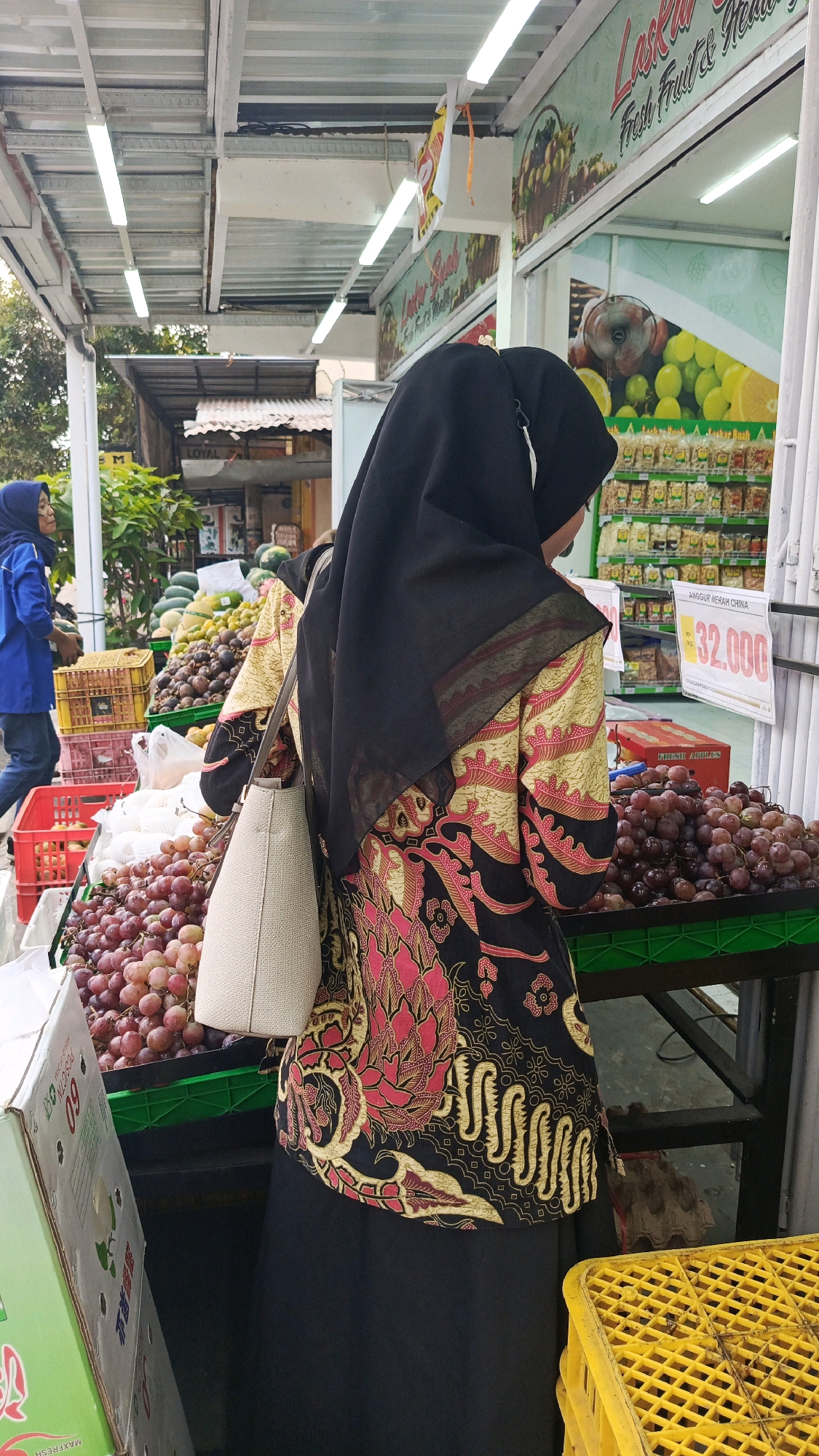 A woman wearing a black hijab and a colorful batik patterned dress is standing in front of a fruit stall at a market in Mangunsuman, Indonesia. She is holding a light-colored handbag. In the background, various fruits such as grapes, watermelons, and mangoes are displayed in crates and on tables. The fruit stall has a green awning and is illuminated by fluorescent lights. A sign above the stall reads "Laskar Buah Fresh Fruit & Healthy". Another sign displays the price "32.000" for "ANGGUR MERAH CHINA". A second woman in a blue jacket and hijab is visible in the far left of the image, carrying a dark object. The scene suggests a typical day at a local Indonesian fruit market.
