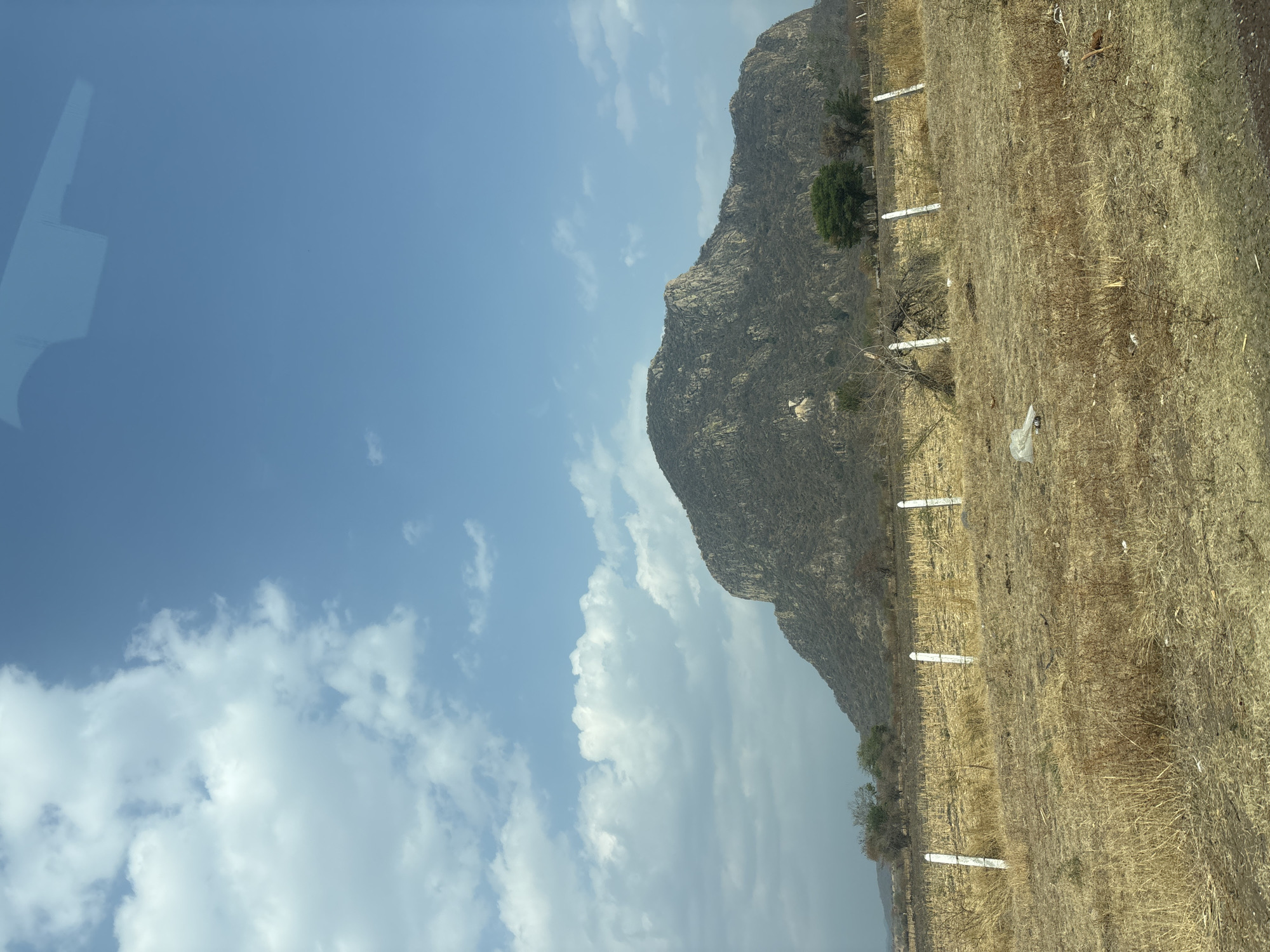 The photograph shows a rocky mountain adjacent to a dry, grassy field. A series of white fence posts run along the border between the mountain's base and the field. The sky is blue with scattered white clouds. The location is within the vicinity of Jantetelco, Mexico.