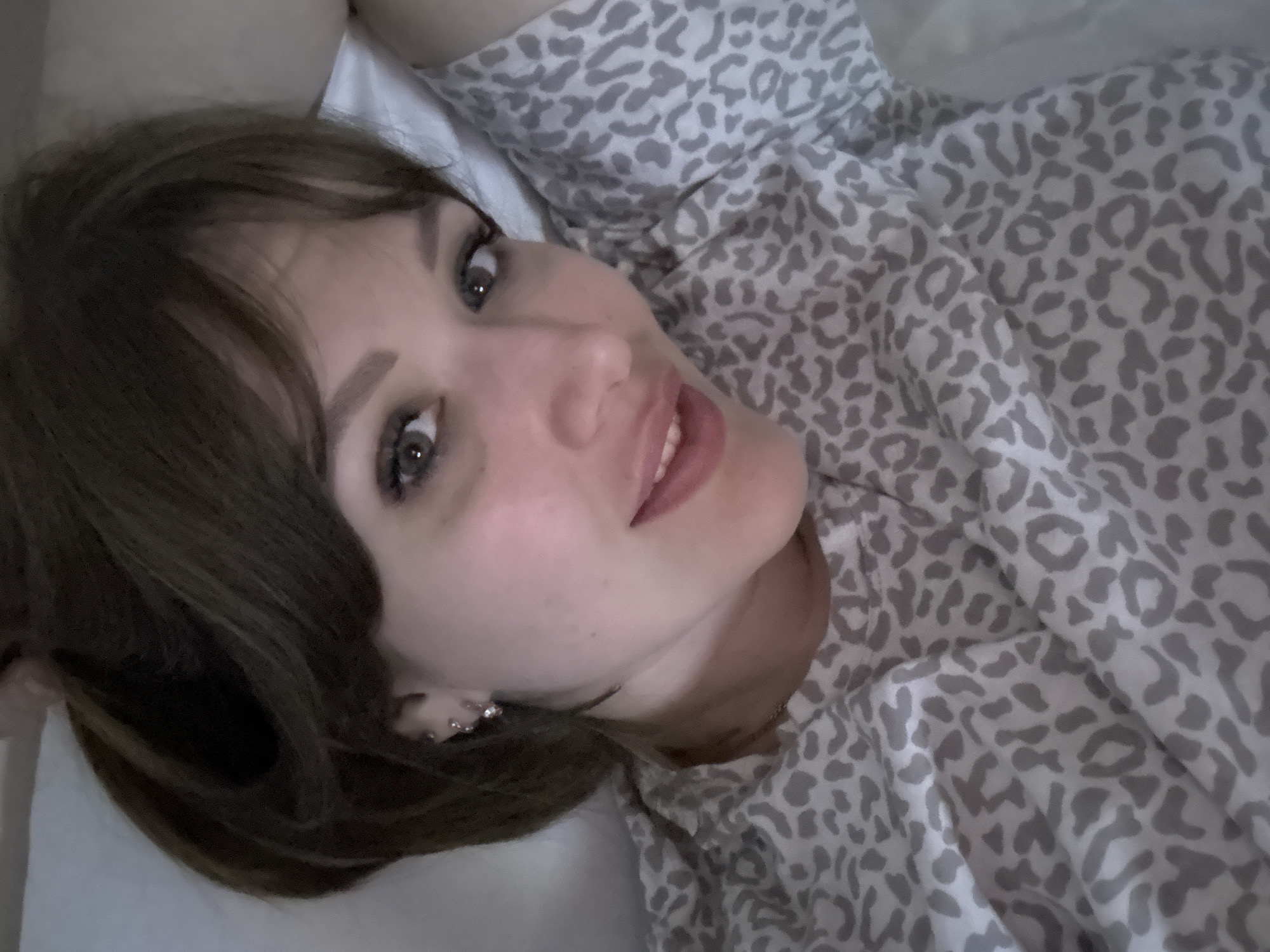 A woman with brown hair and bangs is lying on a white bed. She is wearing a gray and white leopard print top. She is looking directly at the camera. The photo appears to have been taken indoors, possibly in a bedroom. The specific location in Yevpatoriya, Ukraine is not identifiable from the image.
