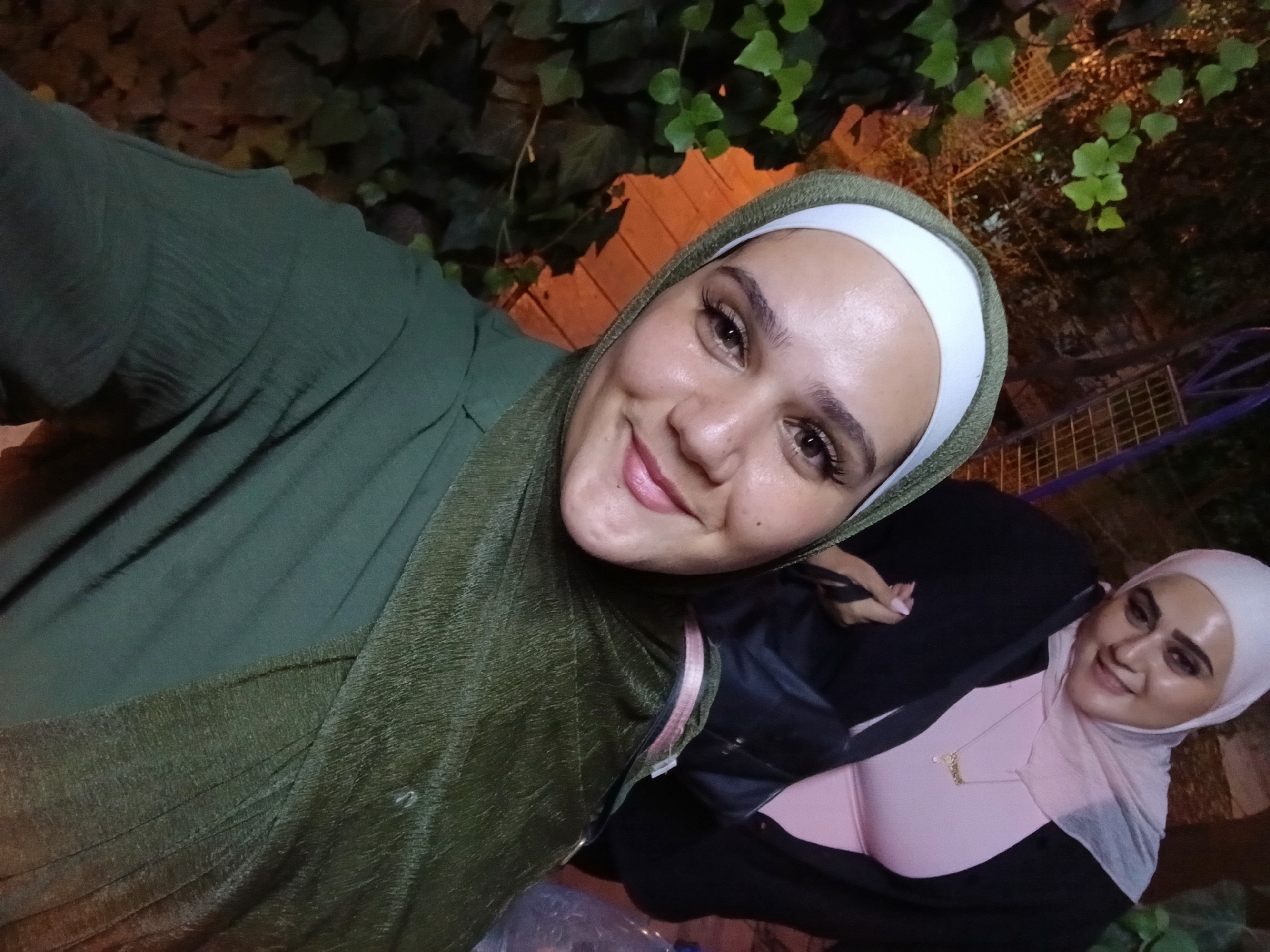 In Qudsaiya, Syria, two women are captured in a selfie taken outdoors at night. The woman in the foreground, wearing a green hijab and a textured green garment, is looking directly at the camera with a smile. Her makeup is visible, with mascara applied to her eyelashes and lipstick on her lips. Behind her, another woman, also wearing a hijab, this one in a pale pink color, is seen smiling. She is wearing a pink top and a black cardigan. A necklace with a pendant is visible around her neck. In the background, there are green plants, a brick wall, and some outdoor lighting. A partially visible plastic bottle is also present.