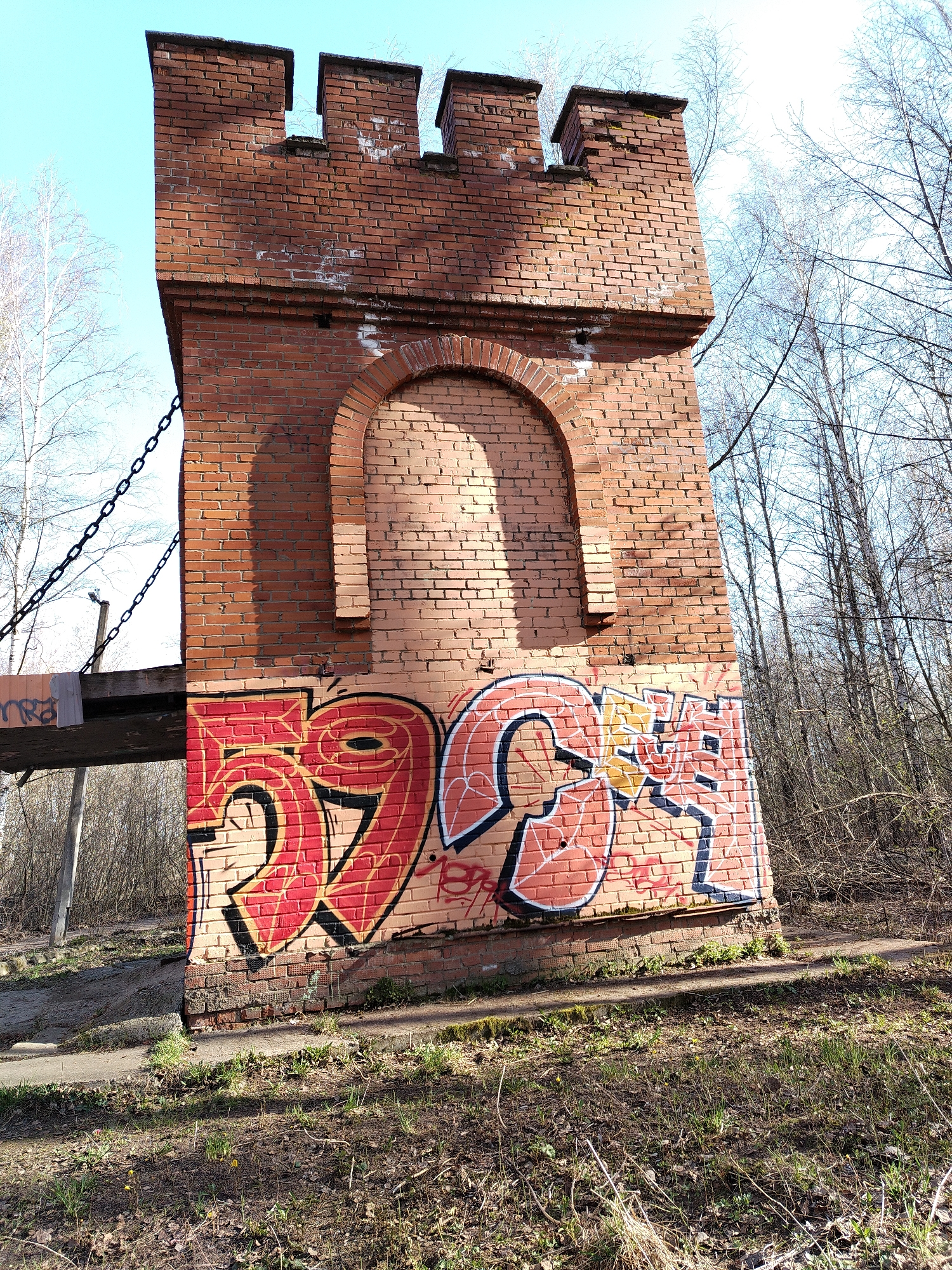 The image depicts a brick tower structure, located in what appears to be a park or wooded area in Chaikovsky, Russia. The tower is rectangular, constructed of red brick, and features crenellations at the top, giving it the appearance of a small castle turret. An archway has been bricked in, located mid-height on one face of the tower. The lower portion of the tower is covered in graffiti, mostly in shades of red, black, and orange, with the numbers 2995 prominently displayed. To the left of the tower, a portion of a bridge or walkway is visible, supported by chains. The surrounding environment consists of trees, some without leaves, and ground covered in grass, dead leaves, and scattered debris. The sky is clear and blue. No people are visible in the frame.