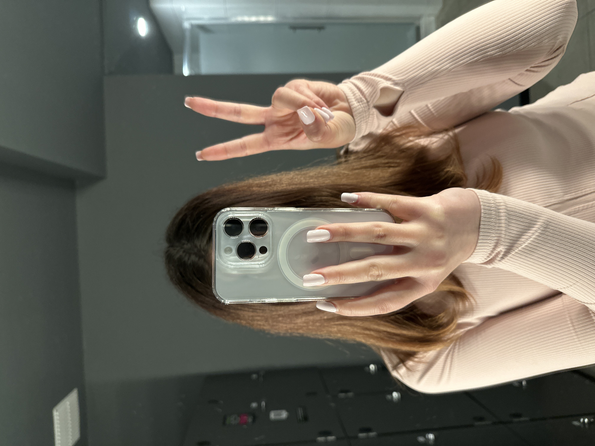 A person is taking a mirror selfie in a locker room. The person is holding a clear phone case with a dark-colored phone inside. They are wearing a light pink, ribbed long-sleeved shirt and have light-colored, square-shaped nails. They are holding up two fingers in a peace sign. The background shows dark gray lockers on the left and a light blue shower stall on the right. The floor appears to be tiled. The image does not contain any specific visual cues that would allow for the identification of the city of Carlow, Ireland.
