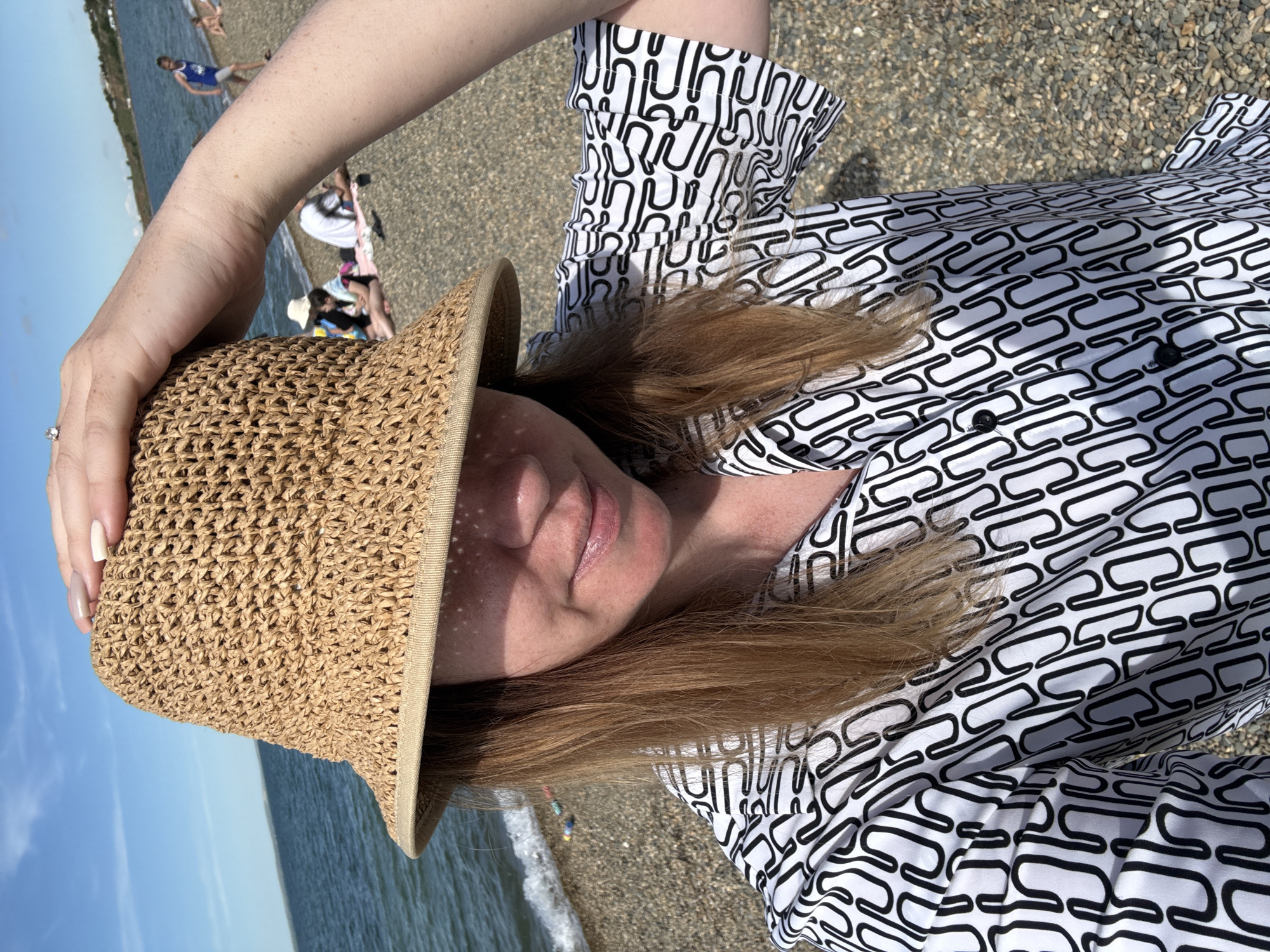 This bright, sunlit image captures a selfie taken by a person on a pebble beach in Taman, Russia. The main subject, seen from a slightly high angle, is holding a light brown, woven straw hat on their head. They have long, light brown hair and are wearing a white shirt adorned with a repeating black pattern of interconnected, elongated 'U' or 'O' shapes. A ring with a small, clear stone is visible on a finger of their left hand, and their nails are painted in neutral shades.
Dappled sunlight filters through the hat, casting intricate shadows across the person's face, highlighting their forehead and nose. A faint, relaxed smile is suggested on their lips.
The background shows a serene beach environment. The shoreline consists of small pebbles or gravel, extending to calm, clear blue water. Beyond the water, the horizon meets a bright, cloudless blue sky. Several other people are present on the beach; one person, possibly a child, in a blue top, is visible standing in the shallow water further along the shore, while a group is seated on the sand closer to the water's edge. Gentle waves can be seen lapping at the shore. The strong sunlight indicates it is likely mid-day or afternoon. No text is visible in the image.