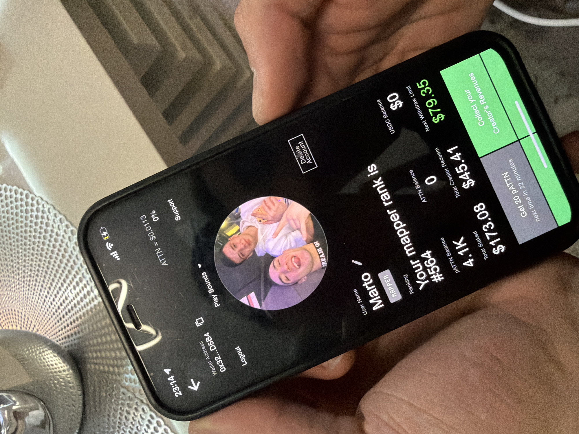 A person is holding a black smartphone, likely an iPhone due to the visible notch, at an angle. The phone's screen displays a dark-themed application interface. The background is a blurry indoor setting, revealing a light-colored surface with a geometric pattern and a metallic, textured object on the left.
The app's screen shows a user profile for "Marto," identified as a "MAPPER," with a profile picture featuring two young males, one smiling broadly with a peace sign and the other subtly smiling. The app displays various financial and ranking details. Prominently featured is "Your mapper rank is #564." Other information includes "PATTN Balance 4.1K," "Total Staked $173.08," "ATTN Balance 0," "Total Creator Redeem $45.41," and "USDC Balance $0." There's a "Next Withdraw Limit" of "$79.35" and a button to "Collect your Creator's Revenues." A timed event shows "Get 20 pATTN next time in 32 minutes." Additional details include a "Wallet Address 0x32...D5B4," an "ATTN" value of "$0.0113," and options like "Play Sounds," "Logout," "Delete Account," and "Support." The phone's status bar indicates the time as 23:14 (11:14 PM), with battery and signal icons visible.