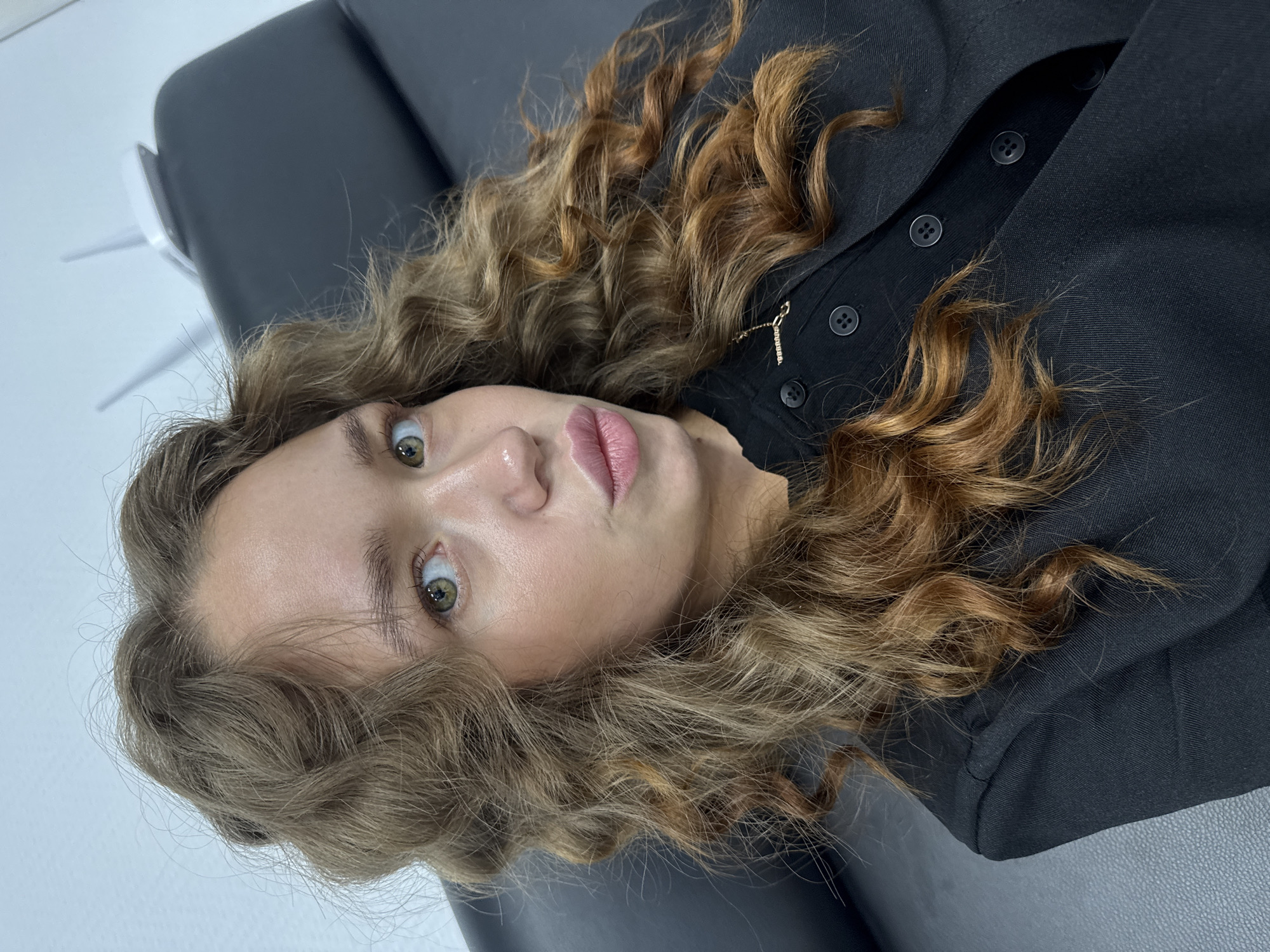 The image shows a young woman with curly, light brown hair, wearing a black collared shirt with buttons and a gold necklace. She is looking at the camera with her lips pursed. She is seated in what appears to be a chair, possibly in an office or clinic setting, indicated by the white wall and a piece of equipment in the background that resembles a router or a medical device. The overall lighting is bright, suggesting an indoor environment. There is no visible information in the image that would allow for the determination of her location within the city of Chaikovsky, Russia.