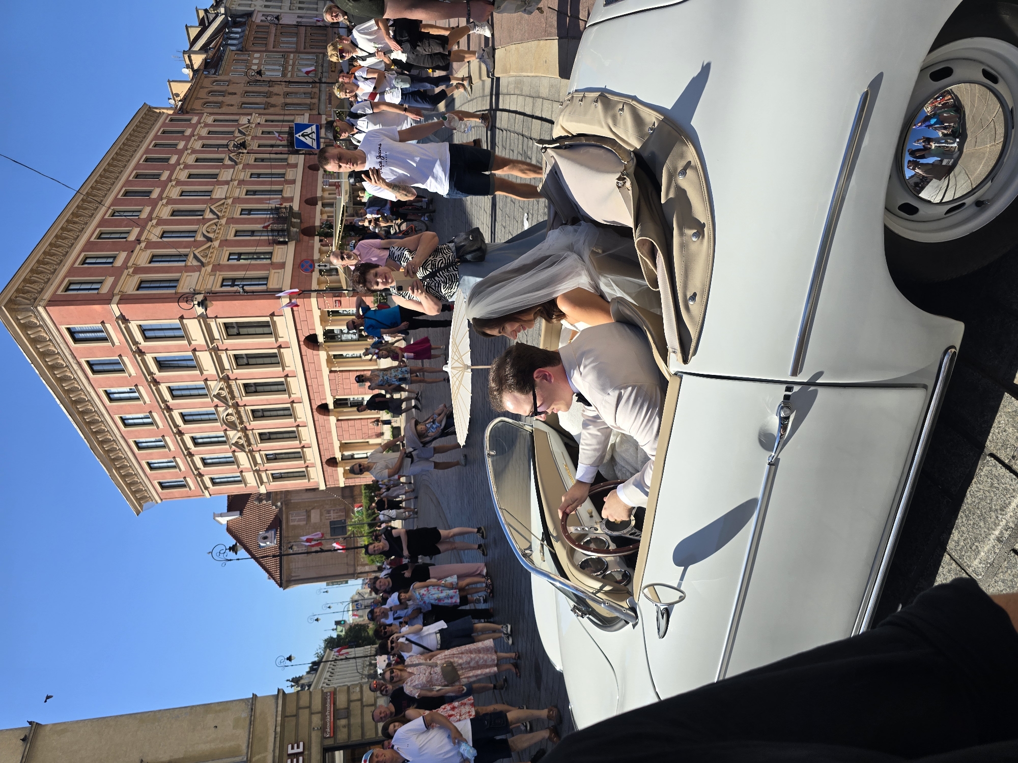 In Warsaw, Poland, a white convertible is parked on a cobblestone street, reflecting a crowd of onlookers. The car, possibly a classic model, has a light-colored interior and is occupied by a man and a woman. The man, wearing a light-colored suit and sunglasses, is seated at the steering wheel. The woman, dressed in a wedding veil and what appears to be a wedding dress, is seated beside him. An umbrella, possibly a parasol, is partially visible. The background features a pink building with multiple floors and windows, characteristic of Warsaw's architecture. The street is lined with people, many of whom are dressed casually and appear to be watching the couple. Some individuals are holding up phones, presumably to take pictures. The sky is clear and blue, indicating sunny weather. The overall scene suggests a wedding celebration or a special event in an urban setting.