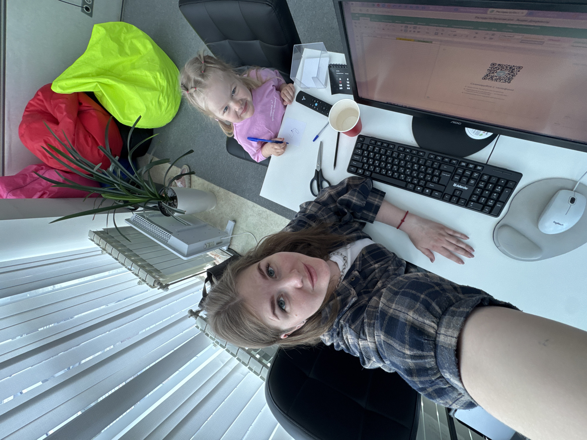 A woman and a young girl are in an office space in Perm, Russia. The woman is in the foreground, facing the camera, with her arms resting on a white desk. She is wearing a plaid shirt and seated in a black office chair. Behind her are closed vertical blinds. The young girl, seated in another black office chair, is drawing on a piece of paper with a blue pen. On the desk, there is a computer monitor displaying a QR code, a black keyboard, a white mouse, a remote control, a white tissue box, a red paper cup, a pair of scissors, and a black pen. In the background, there is a green beanbag chair and a plant. Near the plant, there are red and pink soft bags.