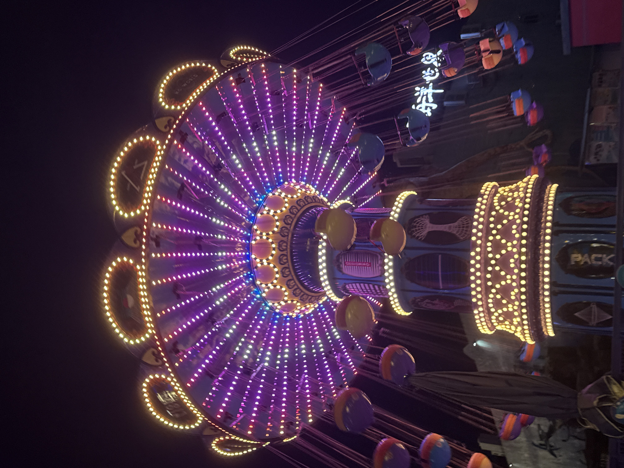 The image shows a brightly lit swing ride at night in the city of Yiwu, China. The ride is adorned with numerous colorful lights, primarily in shades of purple and yellow, creating a vibrant and festive atmosphere. Several swings, some appearing empty and others with unseen occupants, hang from the circular structure of the ride. The background is dark, indicating it's nighttime, with hints of other structures and potentially more attractions in the distance. A neon sign with Chinese characters is visible on the right side of the image. The overall scene depicts a typical amusement park setting, illuminated for evening entertainment.