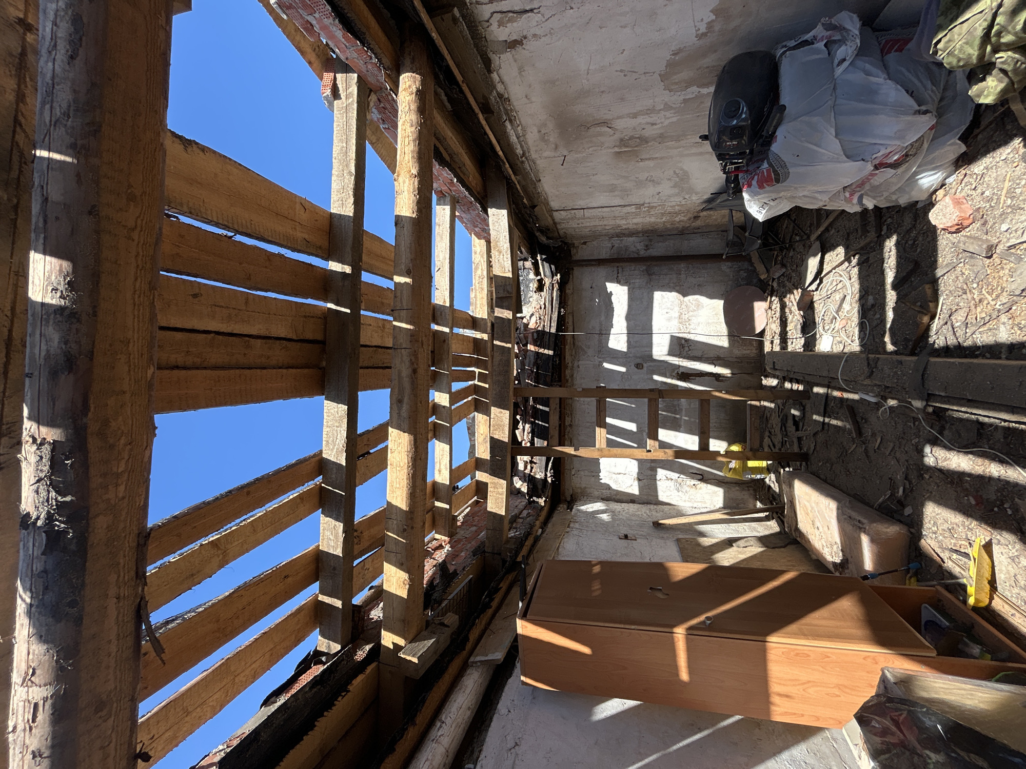 The image depicts the interior of a dilapidated building in what appears to be a state of renovation or demolition. The view is looking upwards towards a partially exposed roof structure, revealing wooden beams and the blue sky beyond. The interior walls are damaged, with crumbling plaster and exposed brick. Debris and construction materials are scattered on the floor, including a wooden ladder, tools, a wooden cabinet, and a bag filled with items. The location of the image is Chaikovsky, Russia.