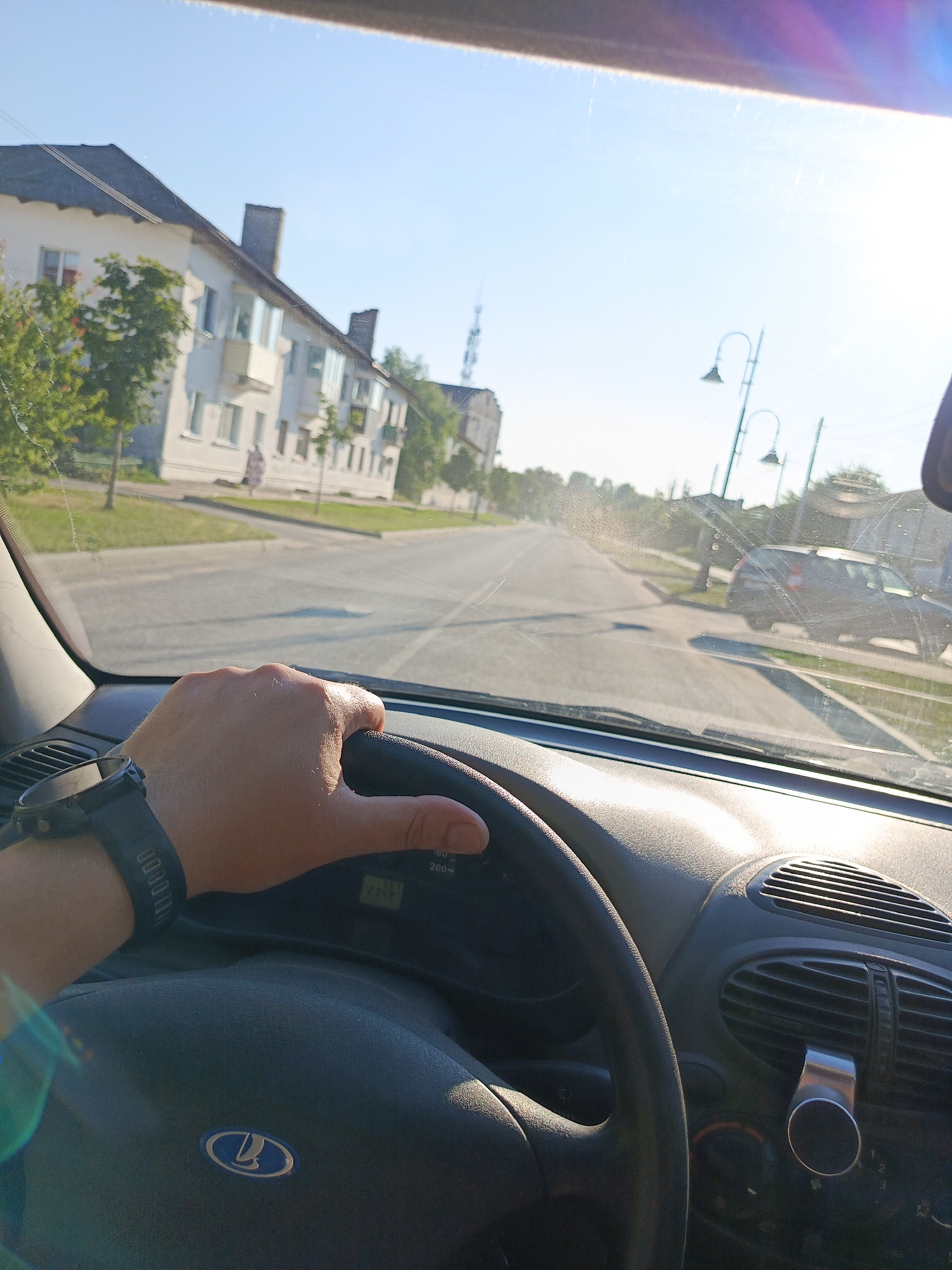 This media file captures a first-person perspective from inside a car, looking forward through the windshield on a sunny day. The driver's hand, adorned with a black smartwatch, rests on the steering wheel. The steering wheel features the Lada logo. The dashboard is visible, with a vent and a metallic phone mount attached to it.
The scene outside the car depicts a residential street lined with multi-story buildings and trees. The buildings appear to be apartment blocks, suggesting an urban or suburban setting. Streetlights are visible along the road. In the distance, another car is parked on the side of the road. The sun is high in the sky, casting shadows and creating a bright, slightly hazy atmosphere. The weather appears to be warm and clear.
The overall impression is that of a casual drive through a town during daylight hours. The clear sky and bright sunlight suggest it is likely midday or early afternoon. The location context provided is Uryupinsk, Russia, which aligns with the architectural style and the presence of a Lada car. No specific emotions are conveyed, but the image evokes a sense of everyday travel. Visible text includes the numbers on the speedometer in the car (e.g., 180, 200) and some unreadable text on the driver's watch.