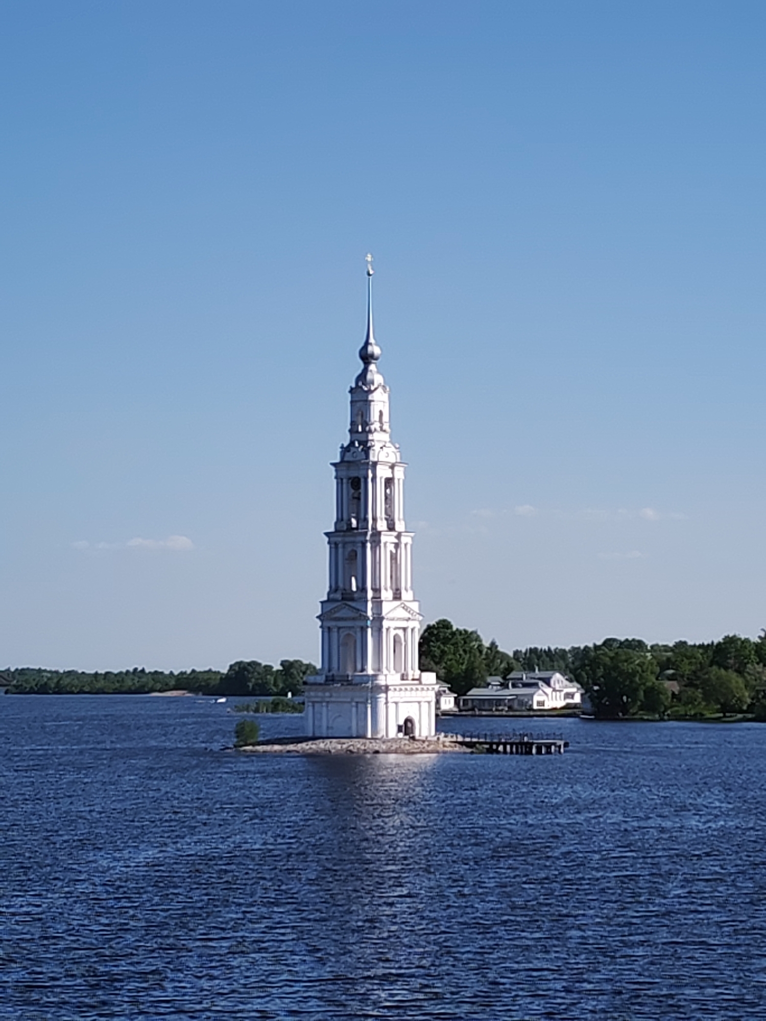 The image shows the Kalyazin Bell Tower, located in Kalyazin, Russia. The white bell tower is situated on a small, partially submerged island in a body of water, presumably the Volga River. The tower is tall and slender, featuring multiple tiers. In the background, there is a shoreline with trees and a white building with a dark roof. A wooden dock extends into the water from the island near the bell tower. The sky is clear and blue, and the water is dark blue with visible ripples.
