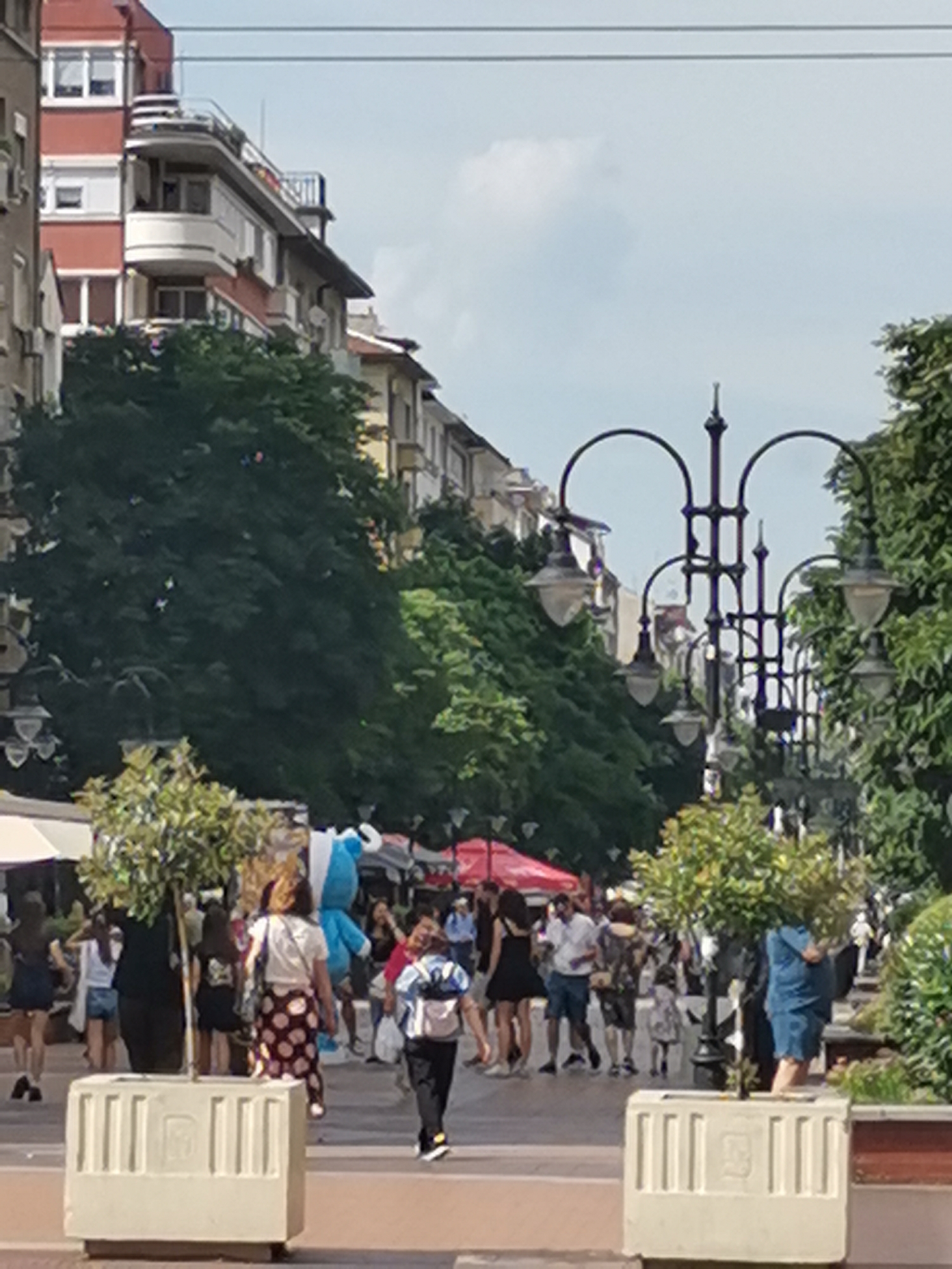 The image depicts a lively pedestrian street in Sofia, Bulgaria, likely on a sunny day. The street is lined with buildings, some appearing older with ornate balconies, and others more modern. Lush green trees are abundant, with some planted in decorative planters. A street lamp with multiple globes stands prominently in the mid-ground.
Several people are walking along the street, engaged in various activities. Some are individuals, while others appear to be in groups or couples. The attire suggests warm weather, with many people wearing casual summer clothing like shorts, dresses, and t-shirts. One person in the foreground is walking with a backpack. In the mid-ground, a person in a blue mascot costume is visible, adding a playful element to the scene. Another person is walking with a large, patterned skirt.
The street itself is paved and appears well-maintained. The overall atmosphere is one of a bustling urban environment, with a mix of architecture and a strong presence of nature through the trees. The image captures a moment of everyday life on a busy thoroughfare in Sofia.