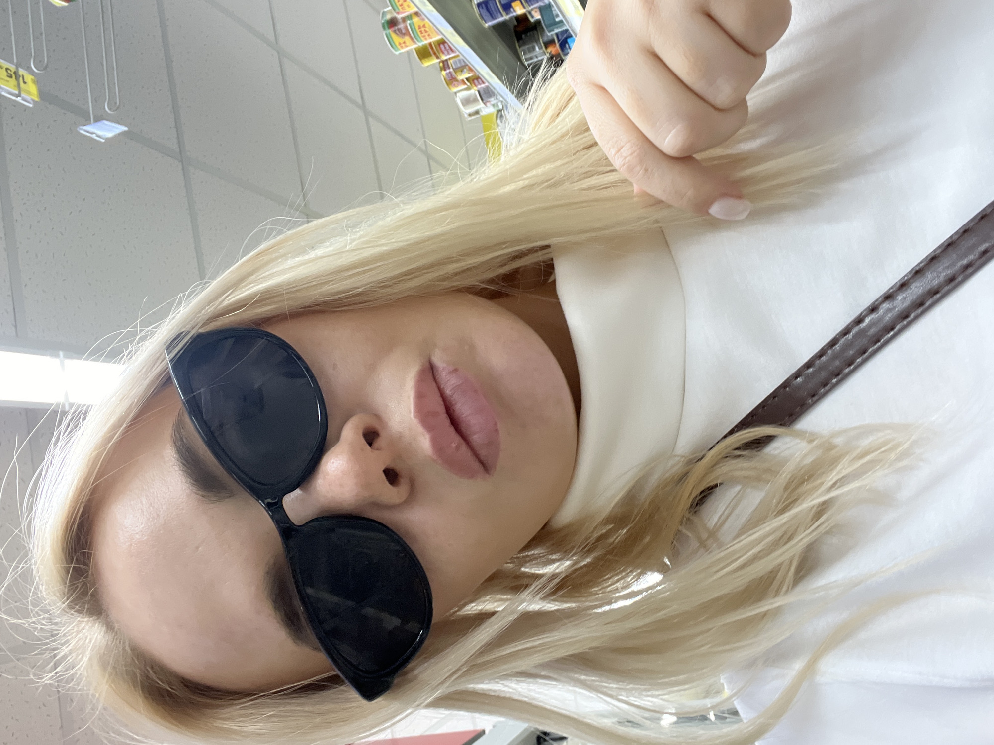 The media file is a close-up, low-angle selfie of a young woman with blonde hair. She is wearing large, black, cat-eye sunglasses and a white top. Her lips are slightly parted, and her expression is neutral. Her hand, with light pink nail polish, is visible near her face, gently touching her hair. A dark brown strap, likely from a purse or bag, is visible across her white top. The background appears to be an indoor setting, possibly a retail environment, with fluorescent lighting and shelves stocked with products visible at the top of the image. There is some text visible on yellow tags in the background, but it is largely illegible. The overall lighting suggests daytime, and the environment appears well-lit. The image is oriented sideways, with the subject's head facing towards the left side of the frame.