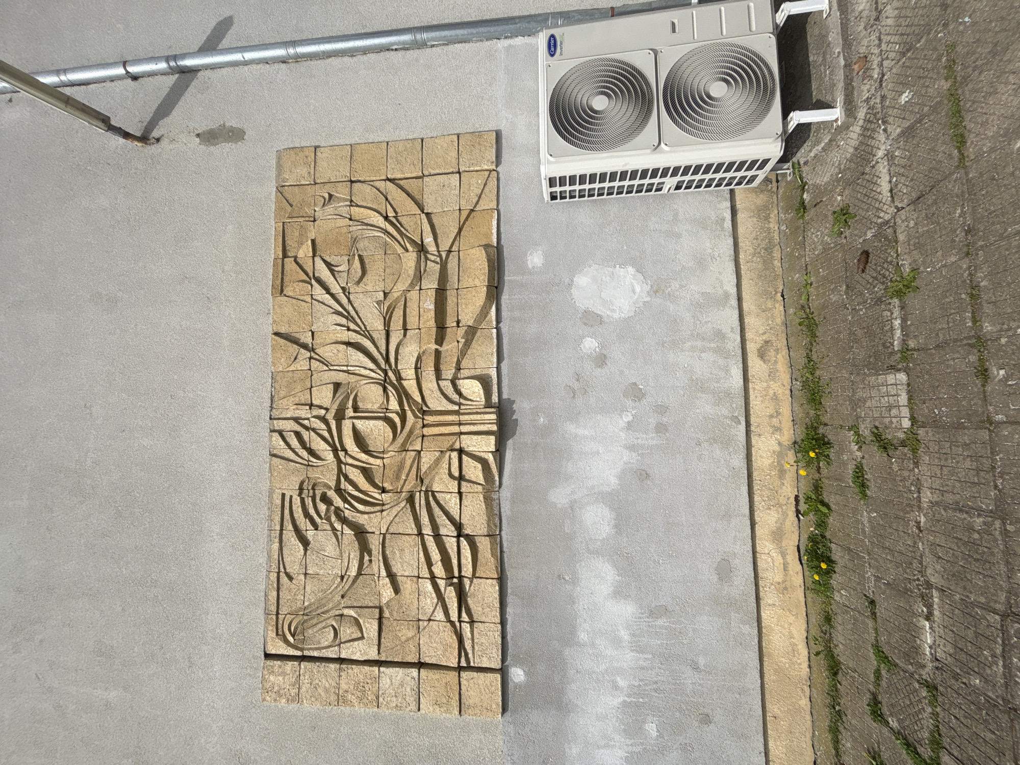 An exterior wall in Troyan, Bulgaria, features a decorative stone relief panel mounted on the wall, depicting an abstract floral design. To the right of the panel, an air conditioning unit is affixed to the wall. A metal pipe runs horizontally near the top of the frame. The wall is painted a light gray color, and a concrete walkway is visible to the right of the wall. Small plants grow in the cracks of the walkway.