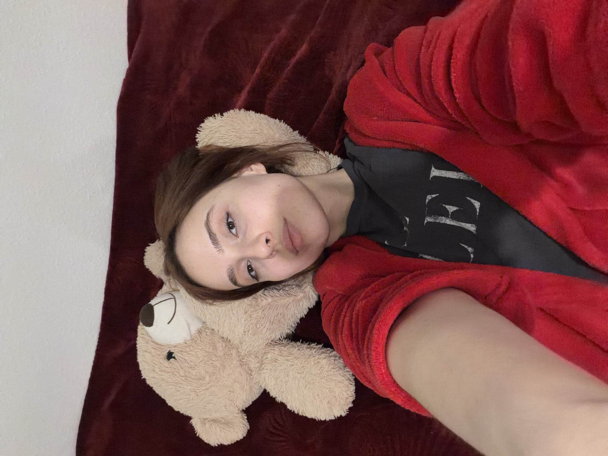The image displays a female subject lying supine on a dark red plush blanket, her head positioned slightly to the right. She has brown hair and is wearing a dark-colored t-shirt with white lettering, partially visible as "LEI", under a bright red fluffy robe. Her gaze is directed towards the camera. Adjacent to her head, on the same blanket, is a light brown teddy bear featuring a dark brown nose and an embroidered smile. The background above the blanket consists of a plain white wall. The scene is an indoor setting in Strasbourg, France.