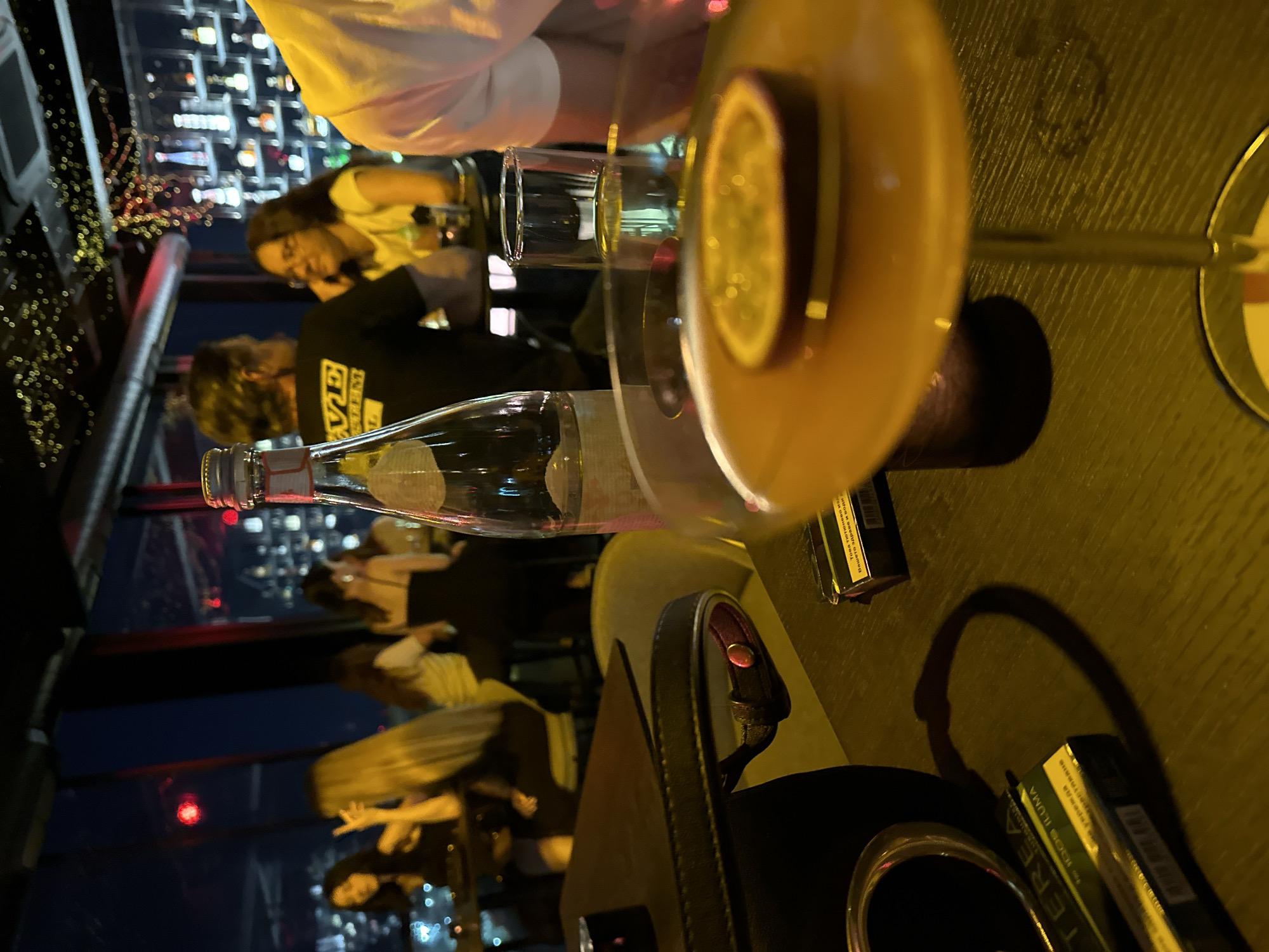 An indoor shot captures a dimly lit bar or restaurant with a panoramic view of a city skyline at night. In the foreground, a table is laden with several items. A cigarette pack, partially obscured, lies on the table. Next to it, a black handbag with a metallic clasp is placed. Dominating the center is a clear glass bottle with a silver cap, standing upright. A cocktail glass, featuring a slice of passion fruit as garnish, is positioned in front of the bottle and slightly to the right. The glass is filled with an amber-colored liquid. In the background, a group of people are seated at tables, their faces blurred. Some are engaged in conversation, while others appear to be looking out at the cityscape. The overall ambiance is sophisticated and relaxed, with soft lighting and subtle decorative lights adorning the ceiling. The location is described as Unknown, Unknown.