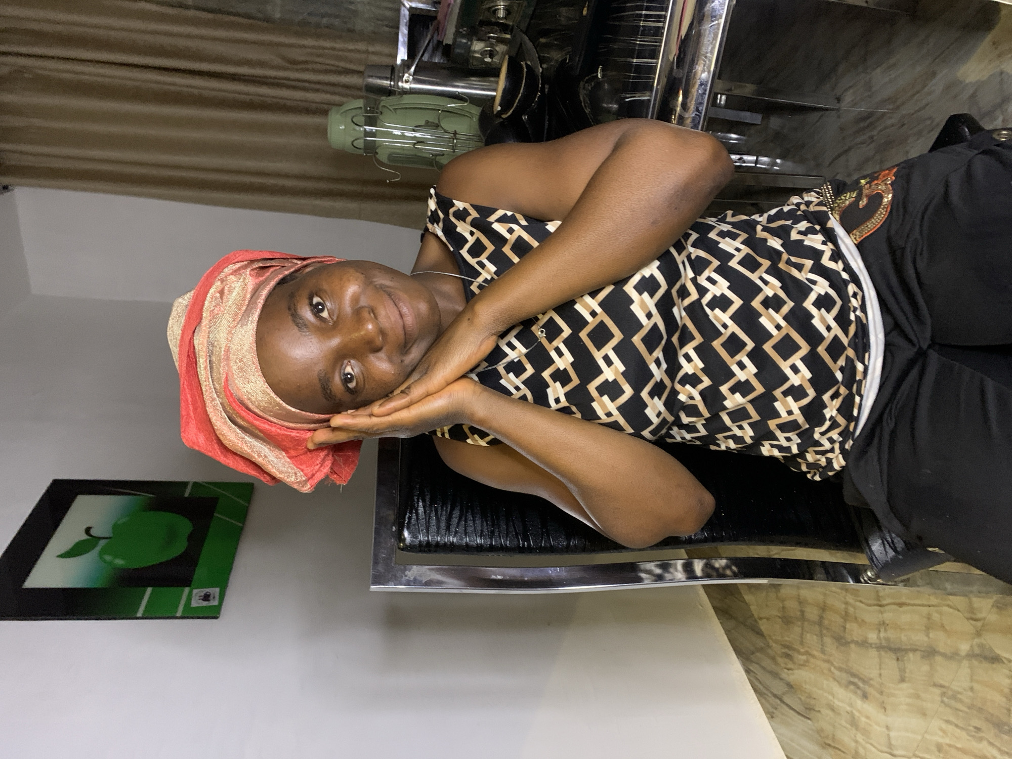 A woman sits in a black chair with a chrome frame. She wears a head wrap of red and gold. She wears a patterned top with black, white, and gold geometric shapes. She is dark-skinned. She is resting her face in her hands and smiling. Behind her is a curtain, a green fan, and what appears to be salon equipment. On the wall is a framed picture of a green apple. The photo was taken indoors in Jos, Nigeria.