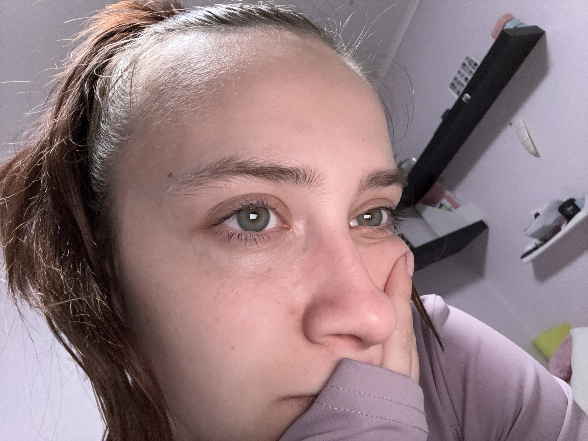 A close-up photograph shows a woman with light skin and brown hair pulled back into a ponytail. Her eyes are green. She is resting her face on her hand. She wears a light purple top. The background features a white wall with a black shelf holding various items including what appears to be sneakers, a book, and other miscellaneous objects. Based on prior information, the location of this image is somewhere in the city of Прикамский, Russia.