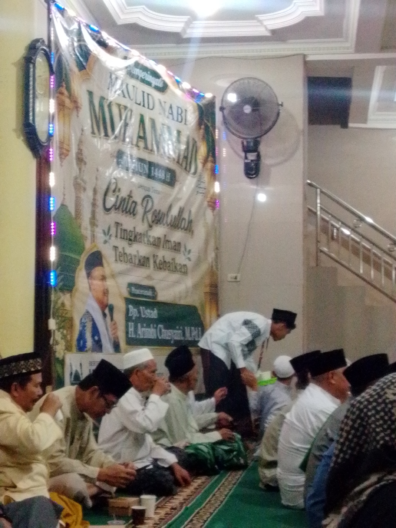 The image depicts an indoor gathering, likely a religious event, at an unspecified location in Bandar Lampung, Indonesia. A large banner hangs prominently in the background, featuring Arabic script and an image of a man speaking into a microphone, identified as "Dyo. Ustad H. Arifin Chuayanni, M.Pd.I." The banner also contains text in Indonesian, including "PERINGATAN MAULID NABI MUHAMMAD" (Commemoration of the Prophet Muhammad's Birthday) and "Tingkatkan Iman, Tebarkan Kebaikan" (Increase Faith, Spread Goodness).
Several men are seated on the floor, facing forward and appearing to listen. They are dressed in traditional attire, predominantly white shirts and various types of caps, including peci. One man in the foreground, wearing a yellow shirt and a patterned cap, is seen interacting with someone to his left. Another man, in a white shirt and peci, is positioned to the right of the banner, appearing to be a speaker or presenter. In the background, a wall-mounted fan is visible, and a staircase with a metal railing ascends to the right. The lighting appears to be artificial, with strings of colorful lights adorning the banner and illuminating the space. The overall atmosphere suggests a community event focused on religious observance.