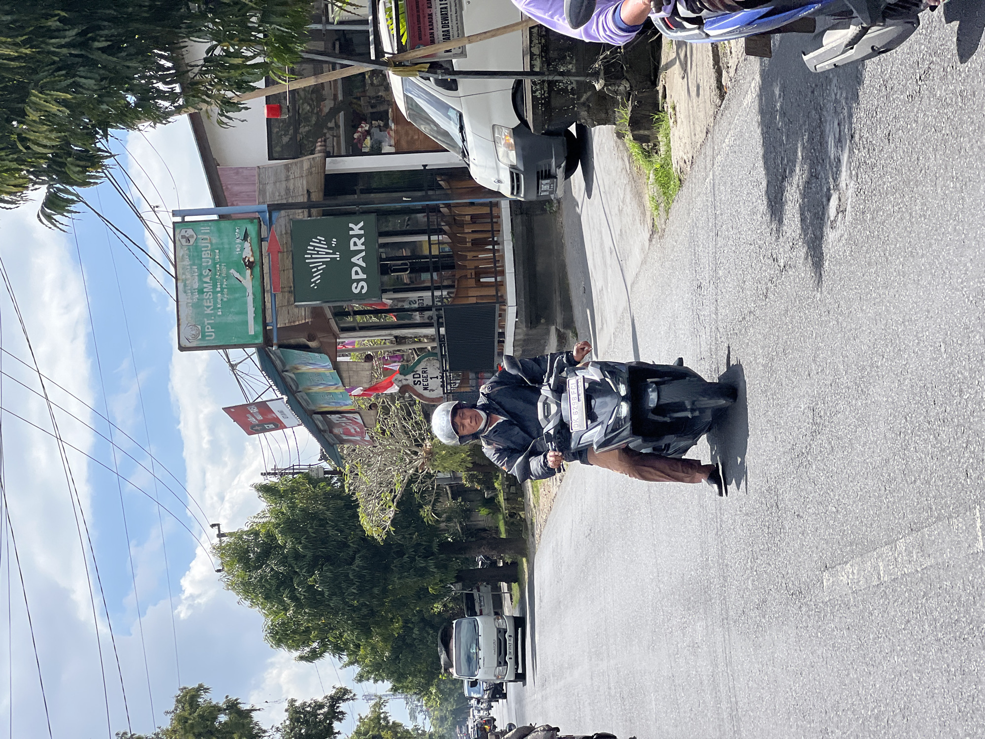 In the city of Ubud, Indonesia, a man wearing a white helmet and a dark jacket rides a black motorcycle down a paved road. He is wearing brown pants and black shoes. The man is looking straight ahead. Behind him, other vehicles, including a white van and more motorcycles, are on the road. The surroundings show buildings with shops, green trees, and a blue sky with white clouds. One building has a sign that reads "UPT KESMAS UBUD II" and another that says "SPARK". The environment appears to be a busy street in a tropical climate.